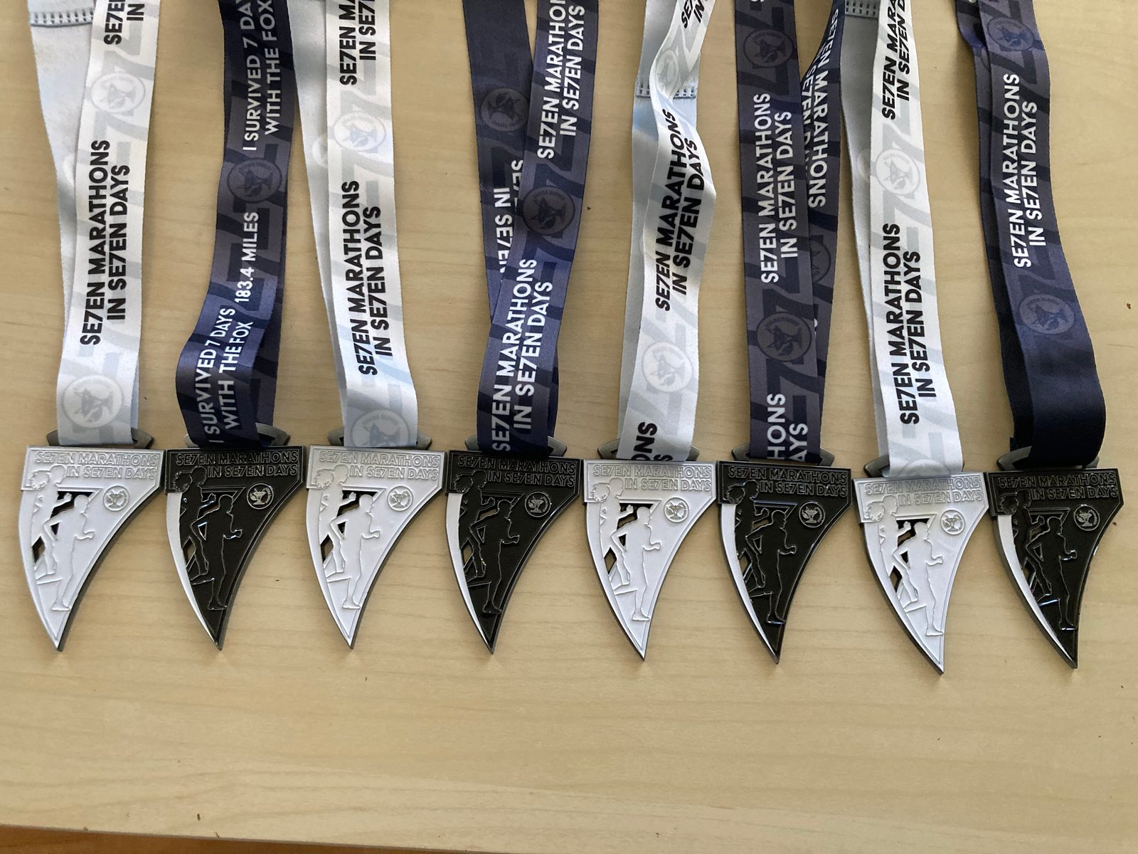 Seven finisher medals with black and white ribbons are laid out in a row on a wooden surface. The medals read “Seven Marathons In Seven Days” and each ribbon features different text related to the event.