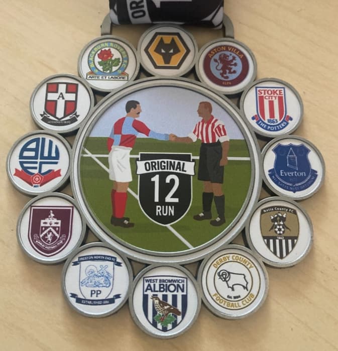 A medal with a center illustration of two soccer players shaking hands on a field, surrounded by 12 soccer club logos. The top band reads "ORIGINAL 12 RUN." The design is on a light wooden surface.