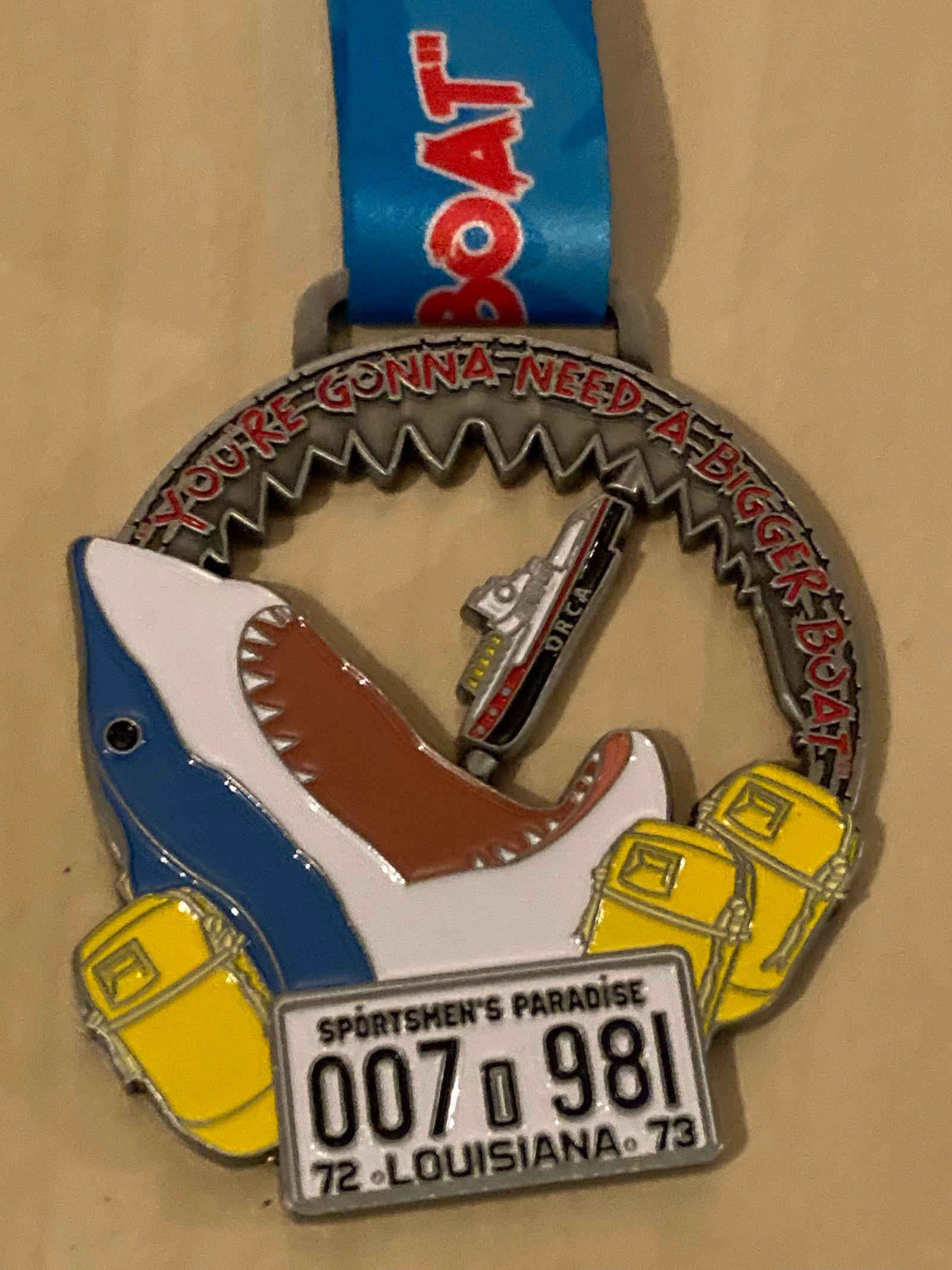 A medal shaped like a shark with its mouth open, featuring a boat inside. It has text at the top saying, "You're gonna need a bigger boat." At the bottom, there's a Louisiana license plate with the numbers "007 981" from 1972-1973.