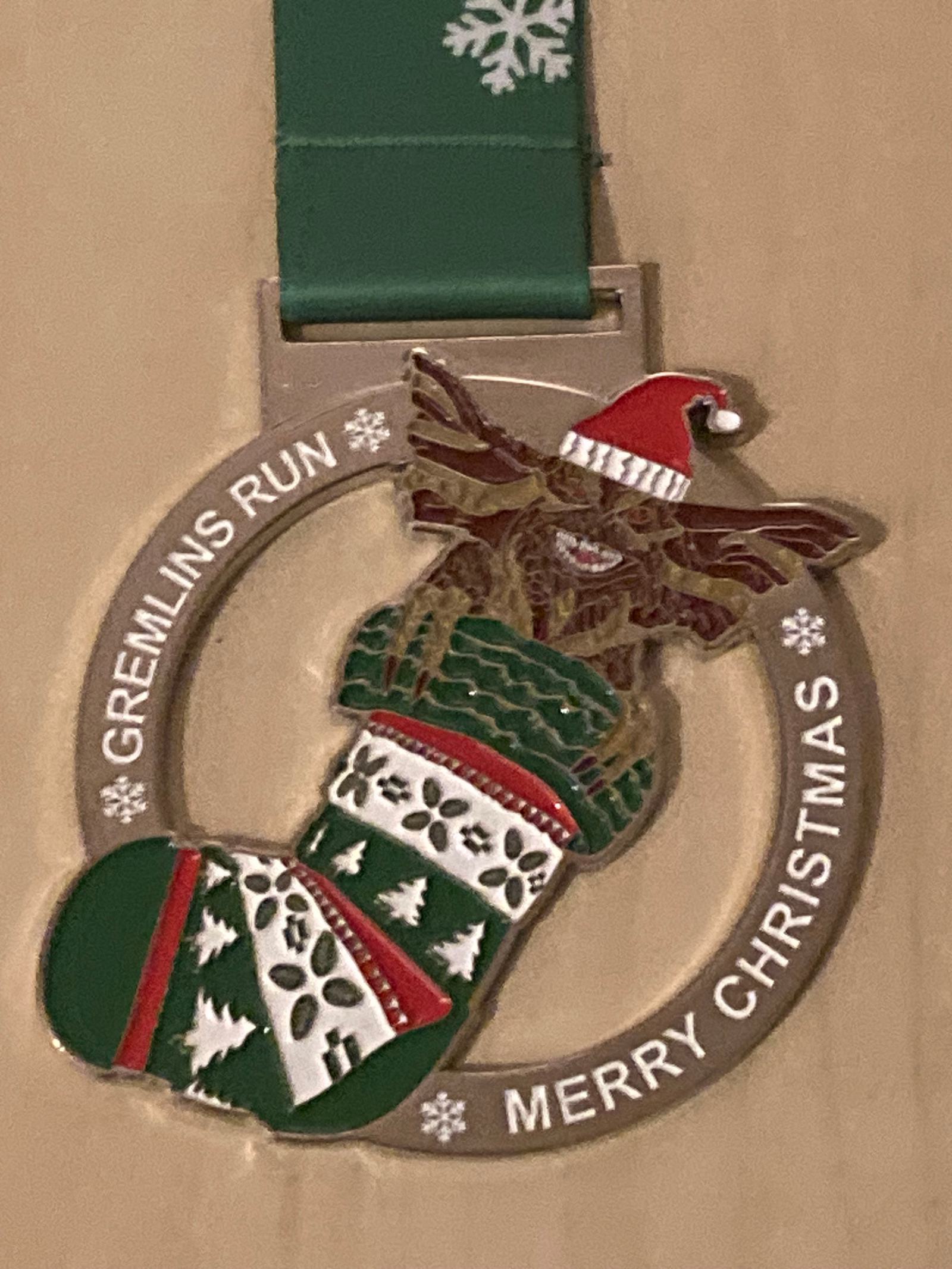 A festive medal featuring a gremlin wearing a Santa hat inside a Christmas stocking with the text "GREMLINS RUN" and "MERRY CHRISTMAS" on the circular frame. The medal is attached to a green ribbon with snowflake designs.