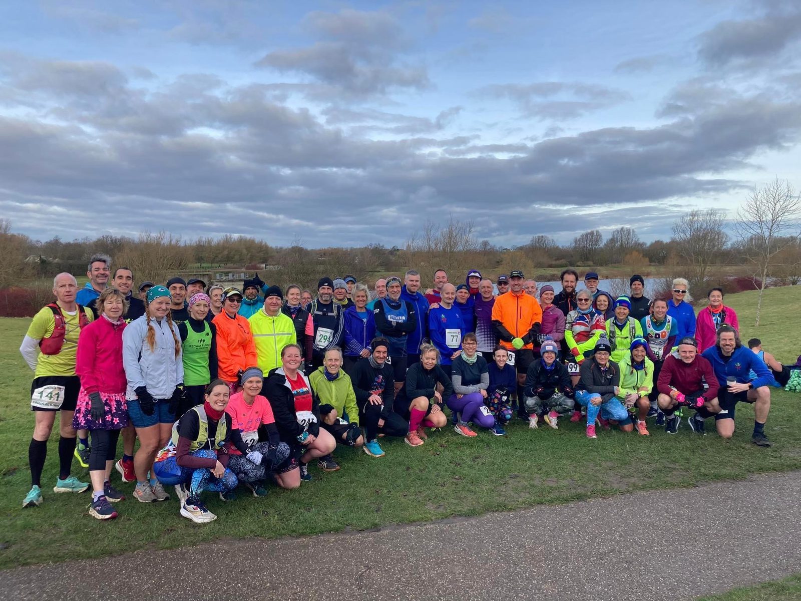 A large group of runners standing and kneeling on a grassy area, posing for a photo. They are dressed in colorful running attire and gear. The sky is overcast with grey clouds. Some runners wear bib numbers, indicating they may be participating in an event.