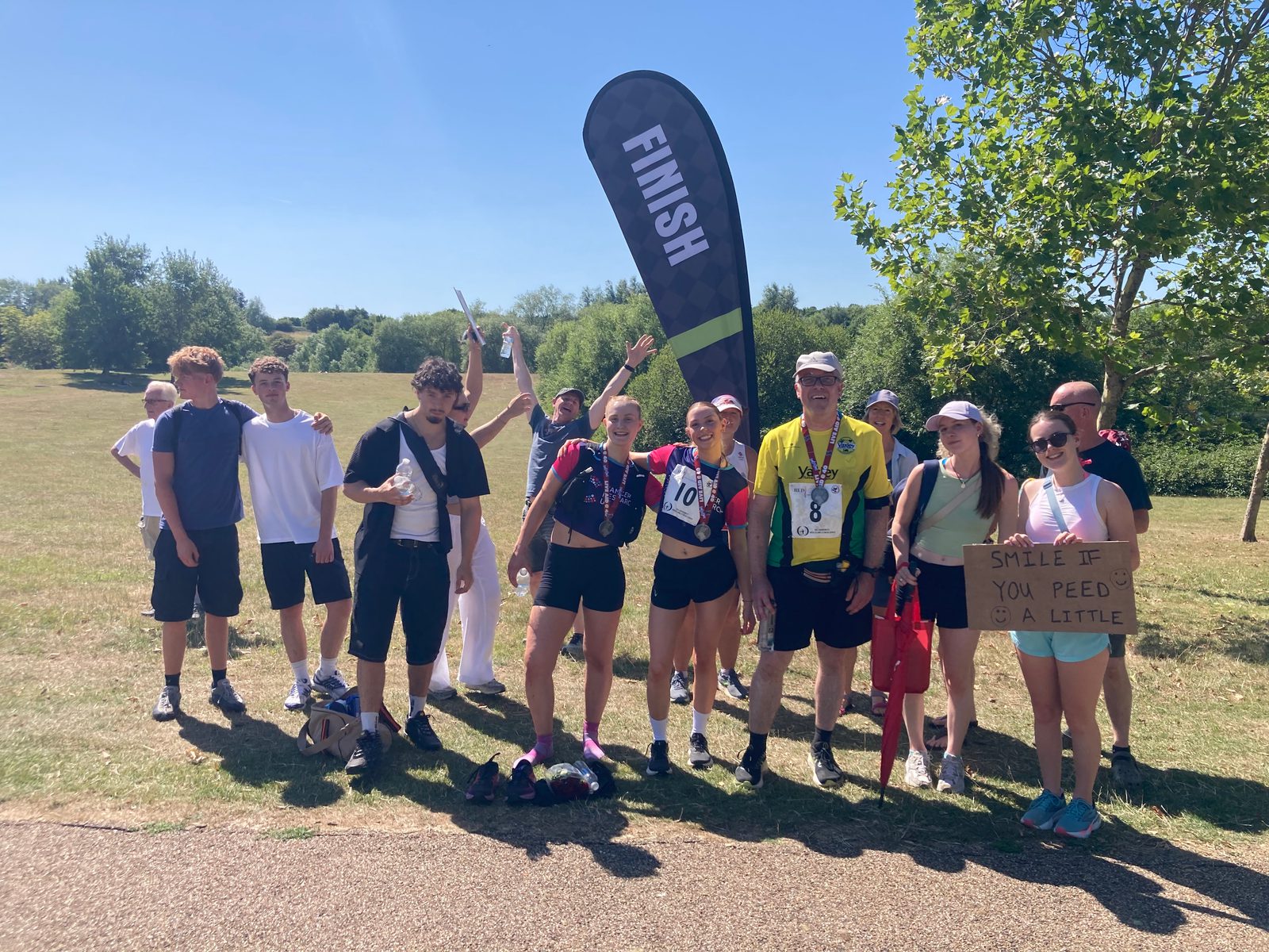 A group of people stand smiling near a "FINISH" flag at an outdoor event. Some wear race numbers, medals, or sports gear. One person holds a humorous sign reading, "Smile if you peed a little." It's a sunny day in a park.