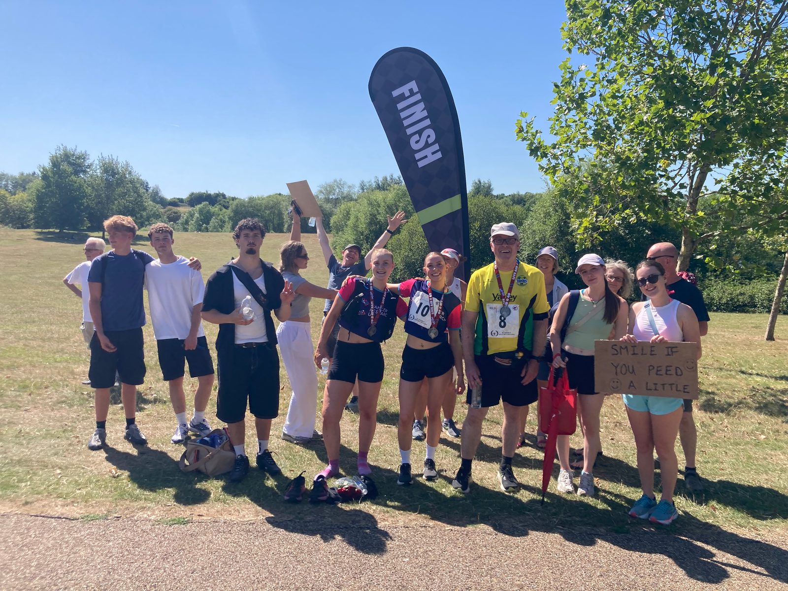 A group of people stands together outdoors in the sun near a "FINISH" flag, some wearing medals and race numbers. Others hold signs, including one reading "SMILE IF YOU PEED A LITTLE." Trees and grass are in the background.