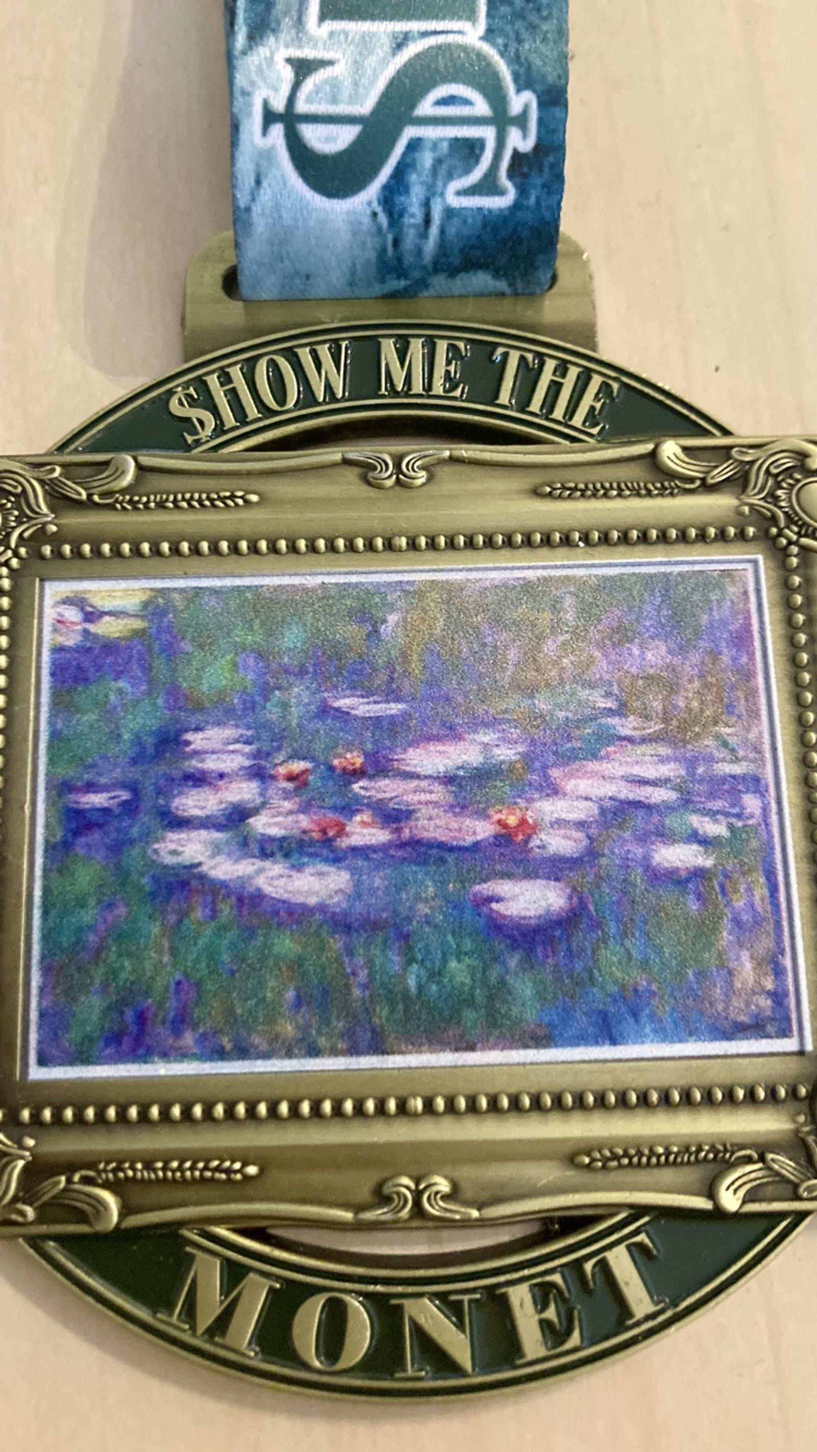 A medal with a painting of water lilies surrounded by green foliage. The text "Show Me the Monet" is engraved above the painting. The medal is attached to a blue ribbon with a white symbol on it.
