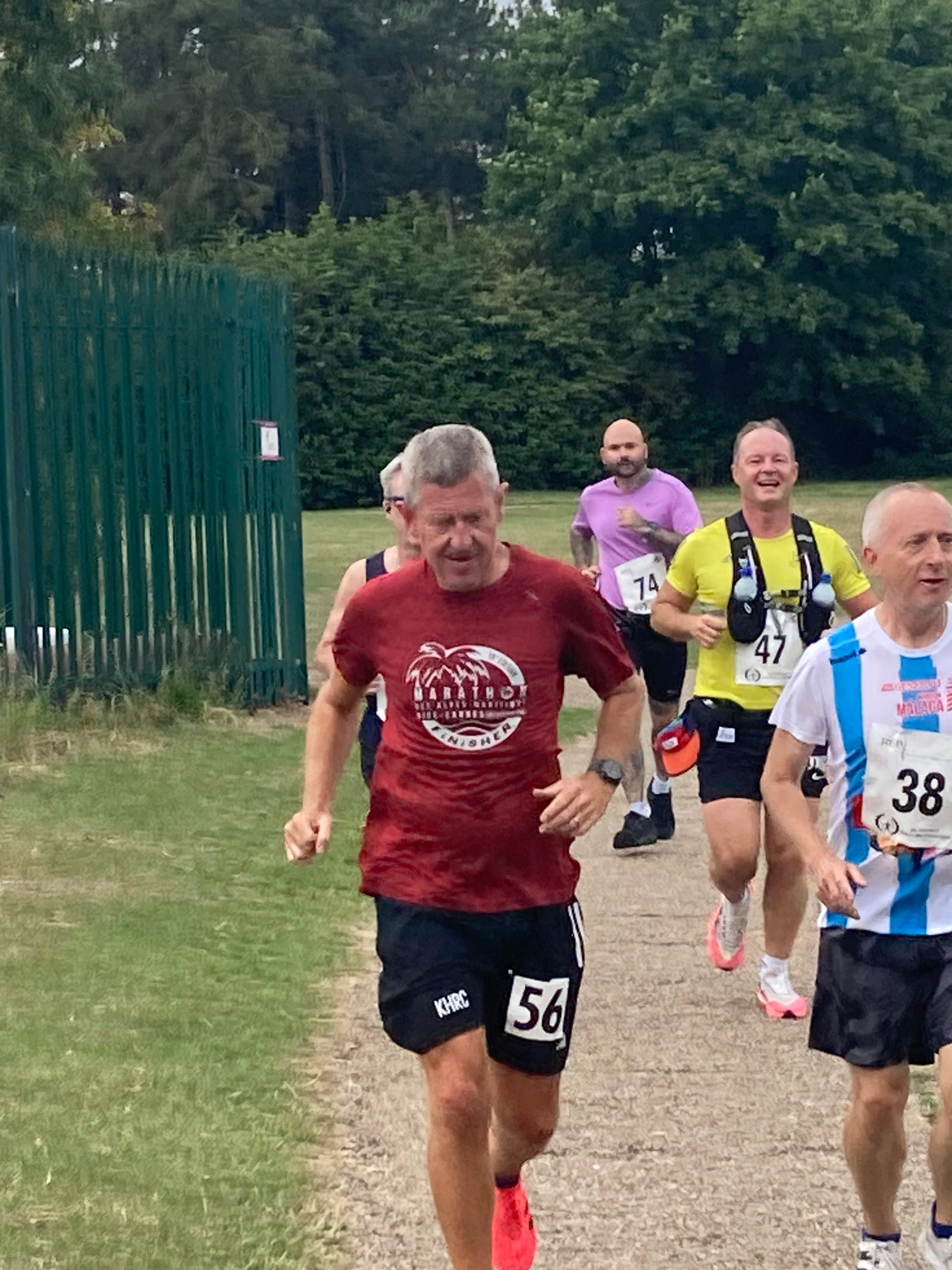 A group of men running a race on a park path, with trees and grass in the background. The man in front wears a red shirt and black shorts with bib number 56. Other runners behind him wear various shirts and race bibs.