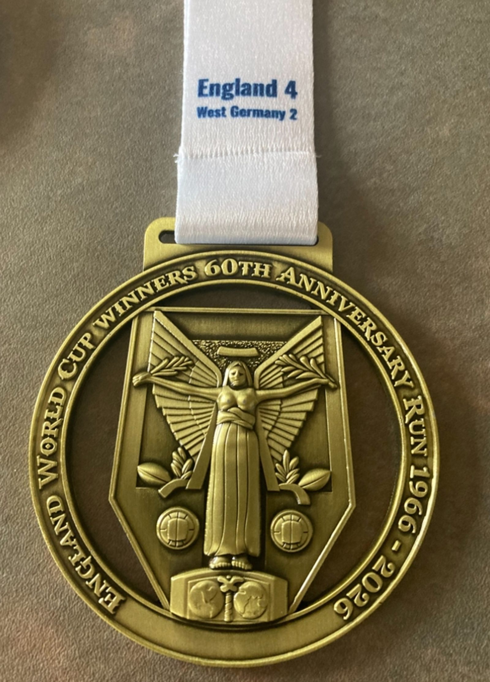 A gold medal with an image of a winged figure and two soccer balls, inscribed "England World Cup Winners 60th Anniversary Run 1966-2026." The ribbon reads "England 4, West Germany 2.