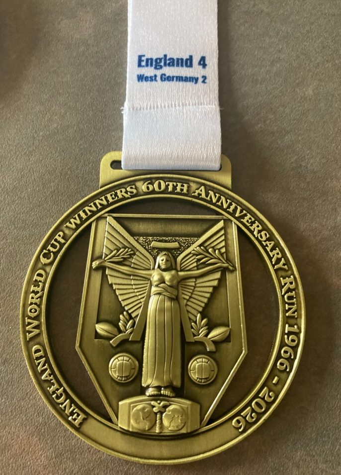 A gold medal with an image of a winged figure and two soccer balls, inscribed "England World Cup Winners 60th Anniversary Run 1966-2026." The ribbon reads "England 4, West Germany 2.