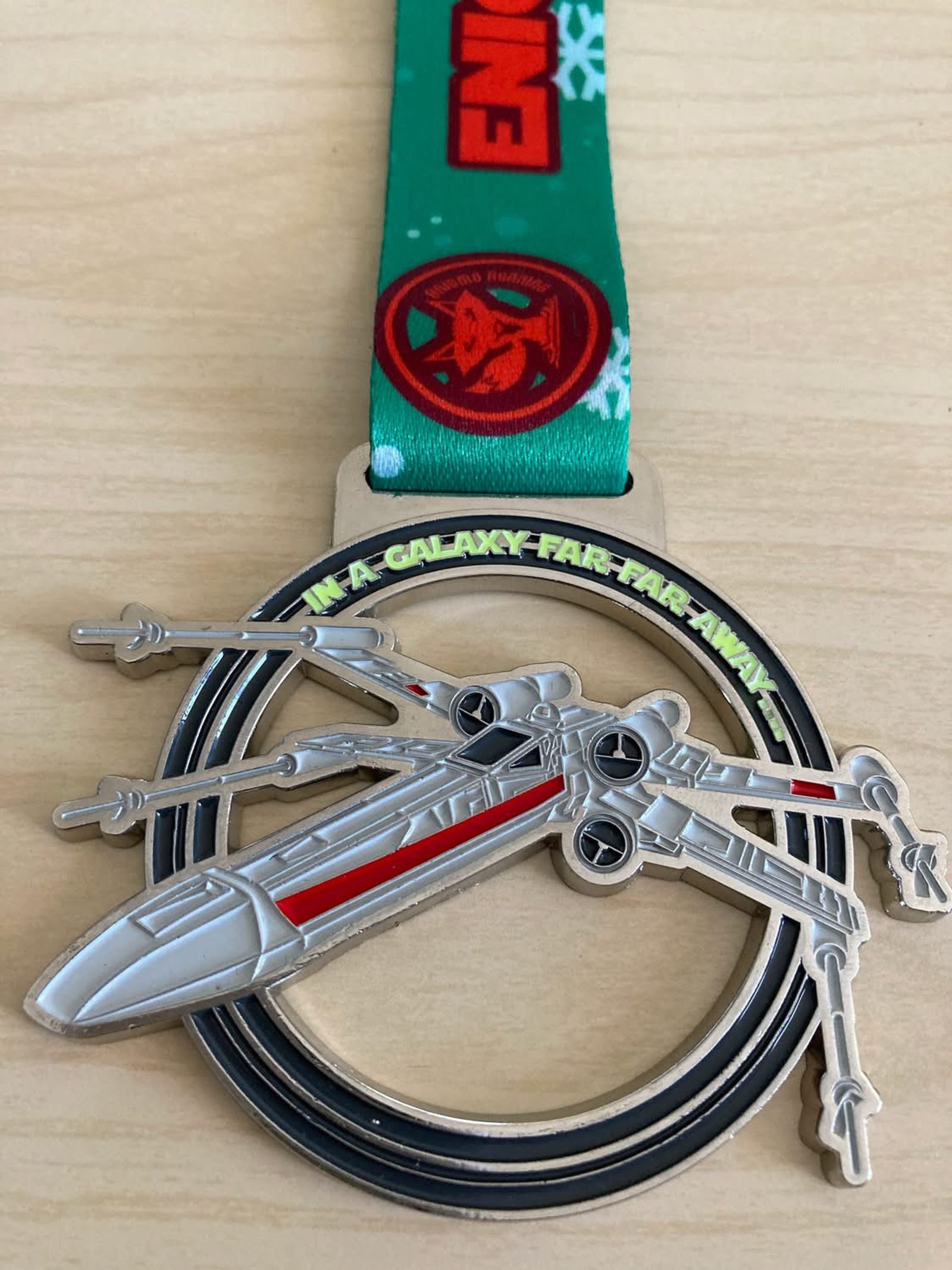 A Star Wars-themed medal featuring an X-wing starfighter and the words "In a galaxy far, far away," attached to a green ribbon with a red emblem, lying on a light wooden surface.