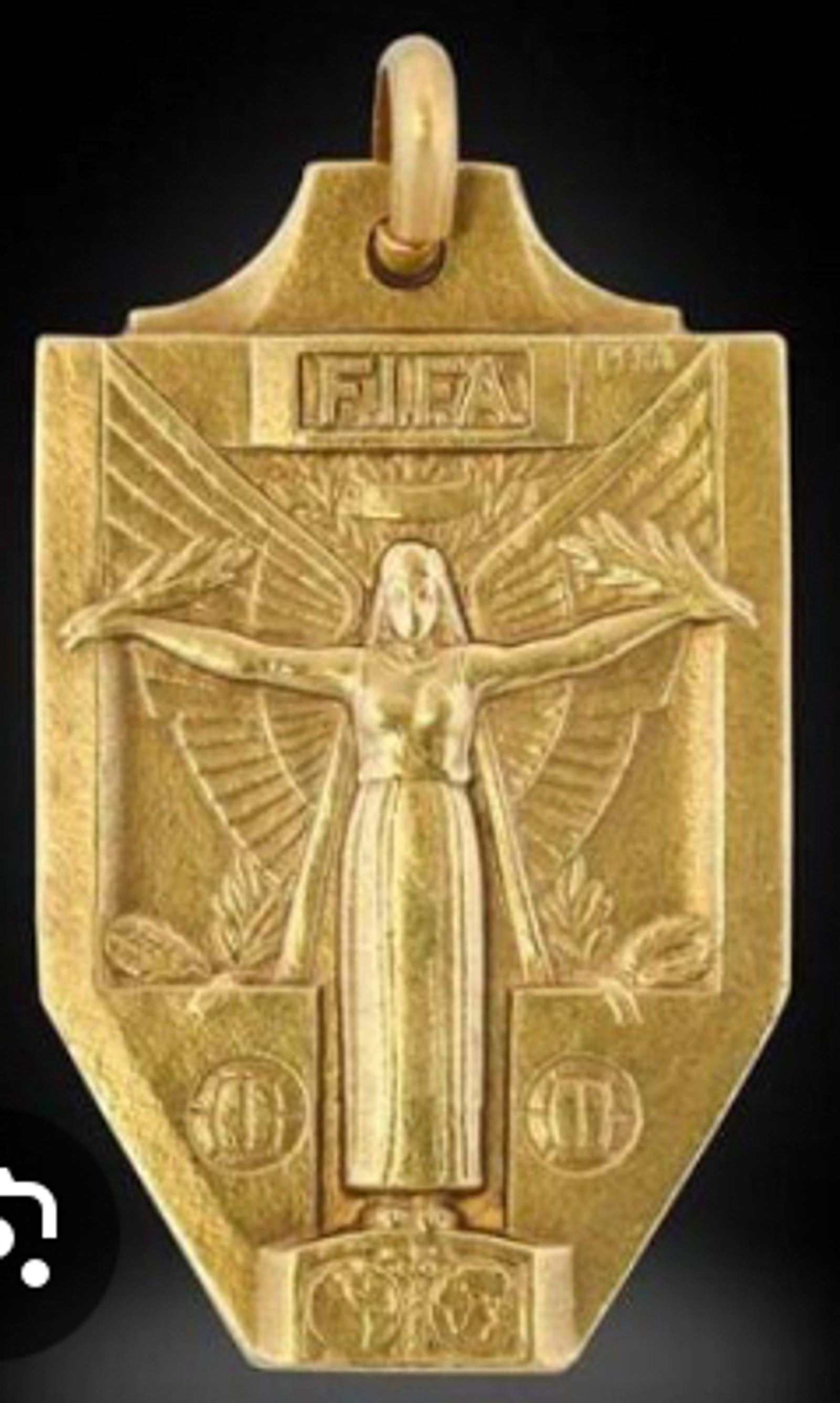 A gold FIFA medallion featuring an embossed figure with outstretched arms and wing-like shapes behind, flanked by circular designs and the "FIFA" inscription at the top.