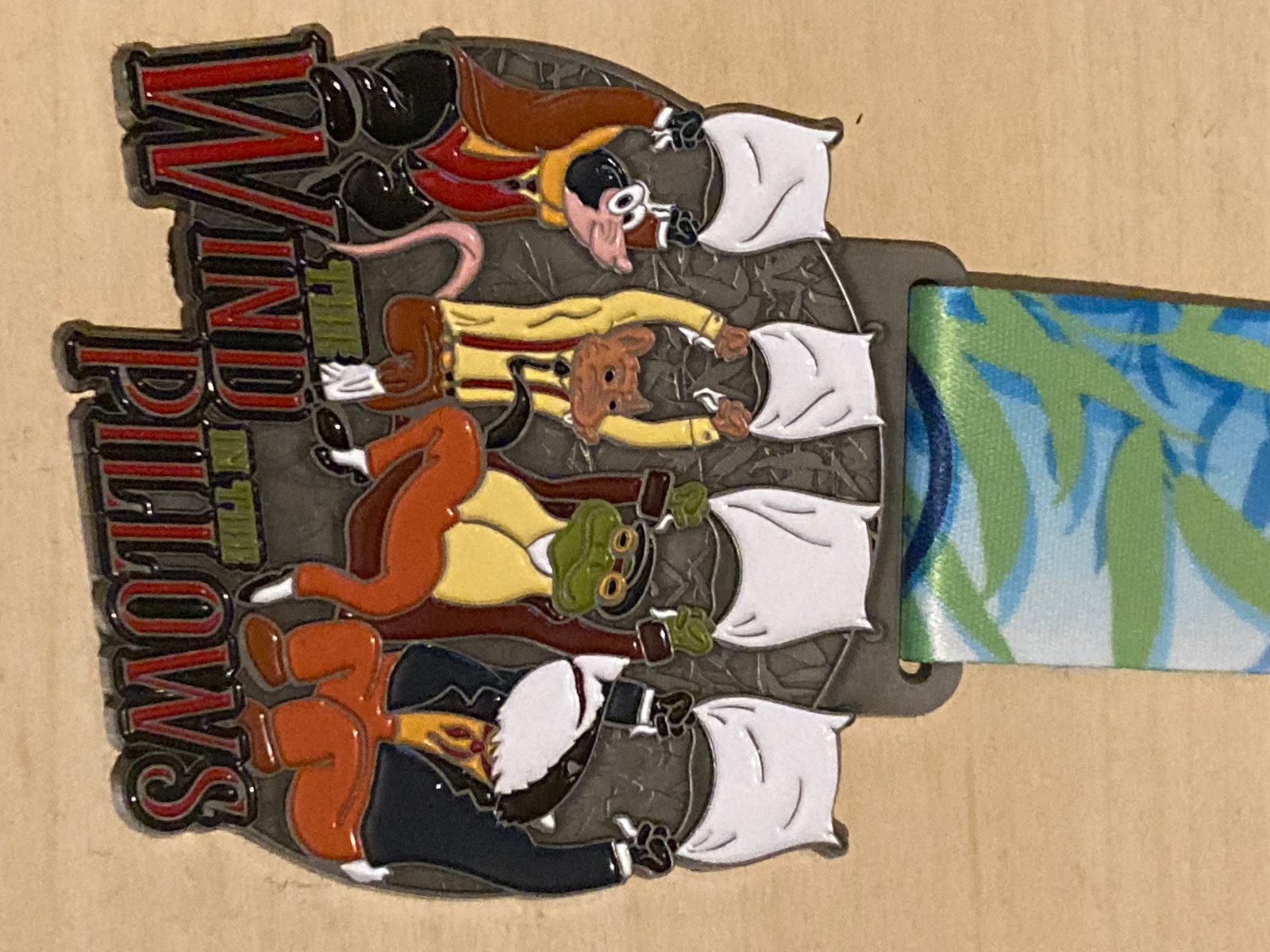 A decorative pin shows four illustrated animals—mole, rat, toad, and badger—holding white pillows, with the text "Wind in the Pillows" in red and green on the left. A blue and green ribbon is attached.