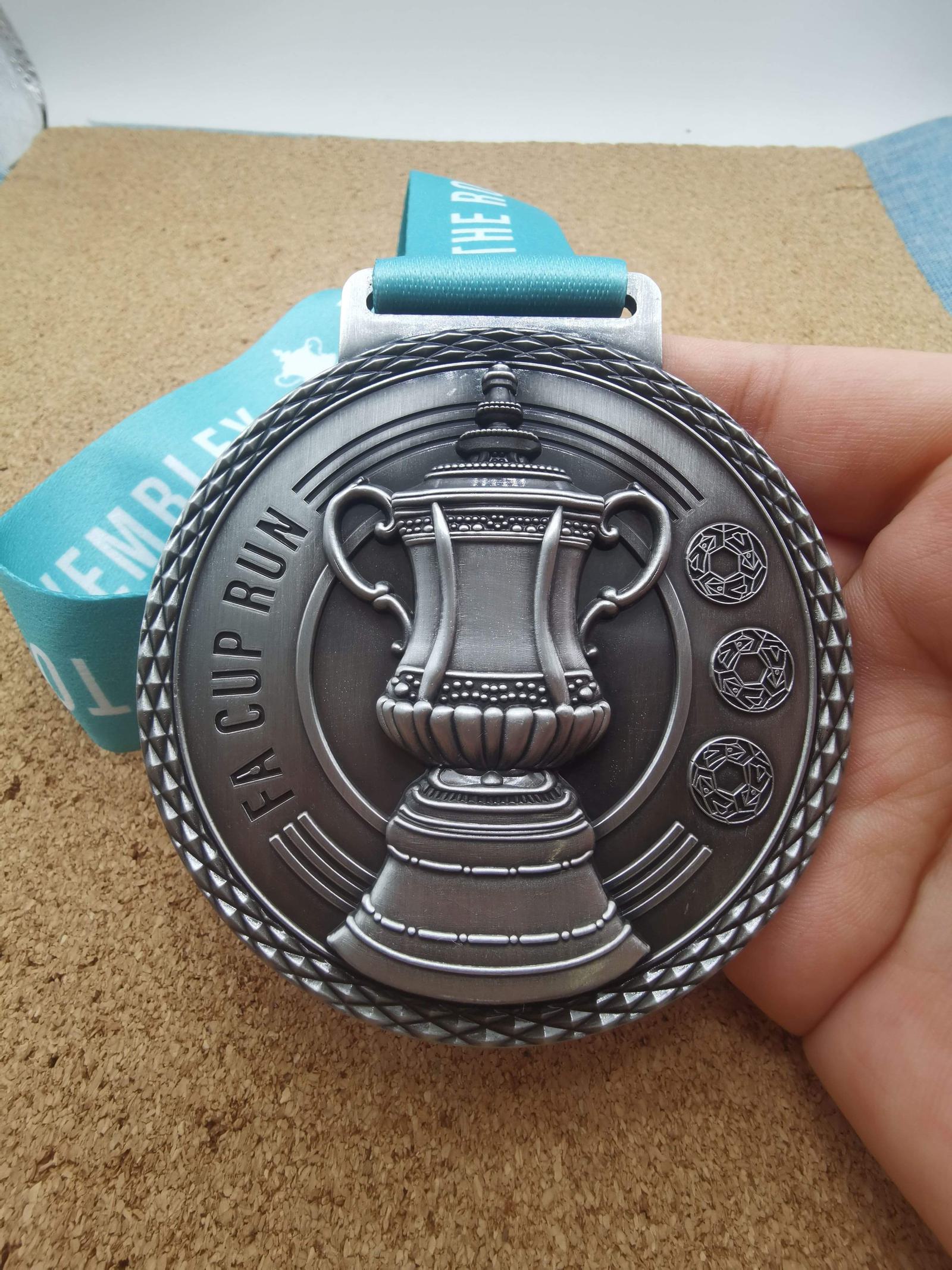 A silver medal with an embossed FA Cup trophy design and text "FA Cup Run." It features three football icons on the right and is held by a hand. The medal hangs from a teal ribbon.