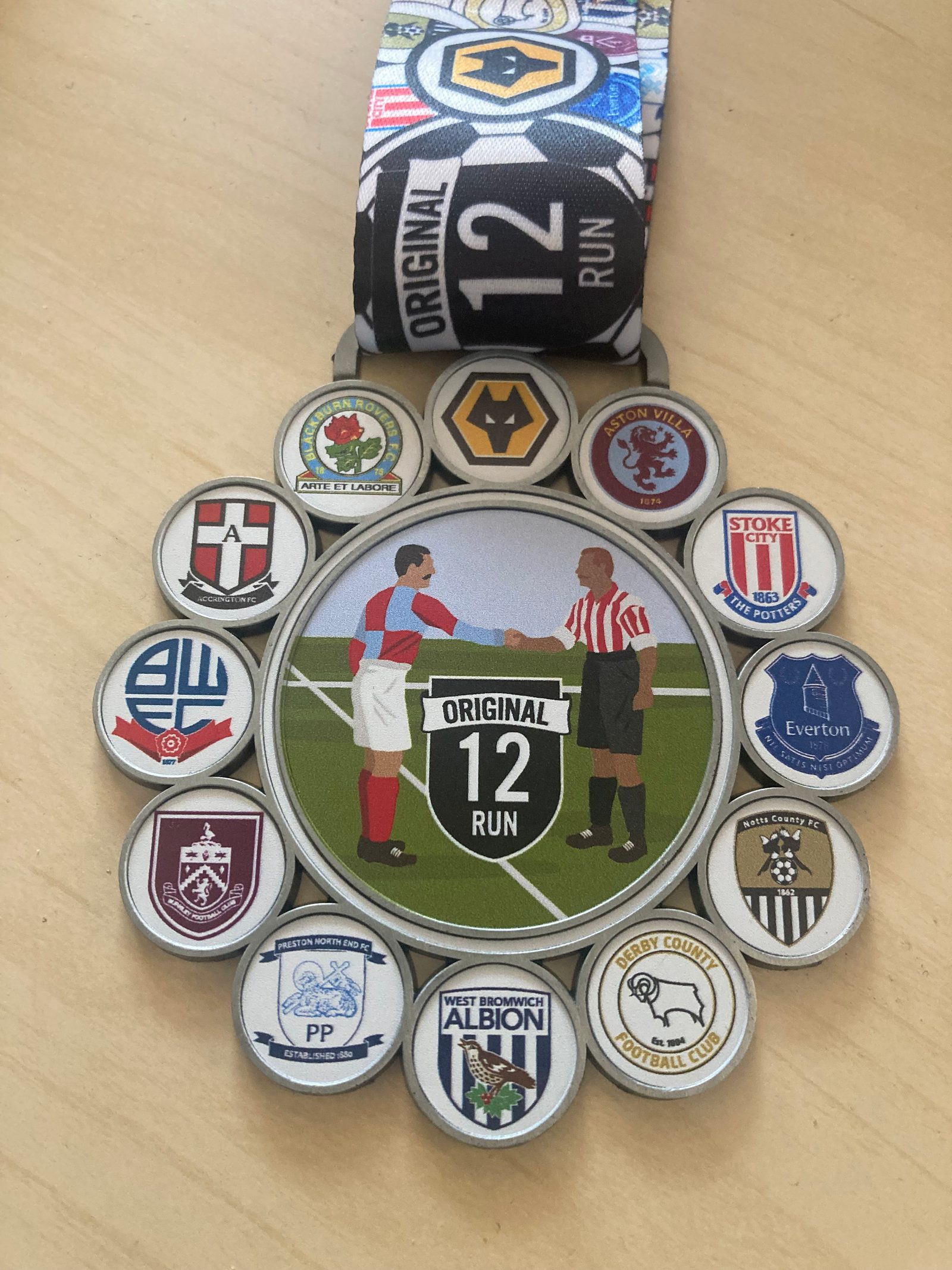 A medal with a central illustration of two football players shaking hands on a field with a "Original 12 Run" logo. Surrounding the illustration are badges of historic English football clubs including Wolves, Aston Villa, Stoke City, Everton, and others. The ribbon is branded "Original 12 Run".