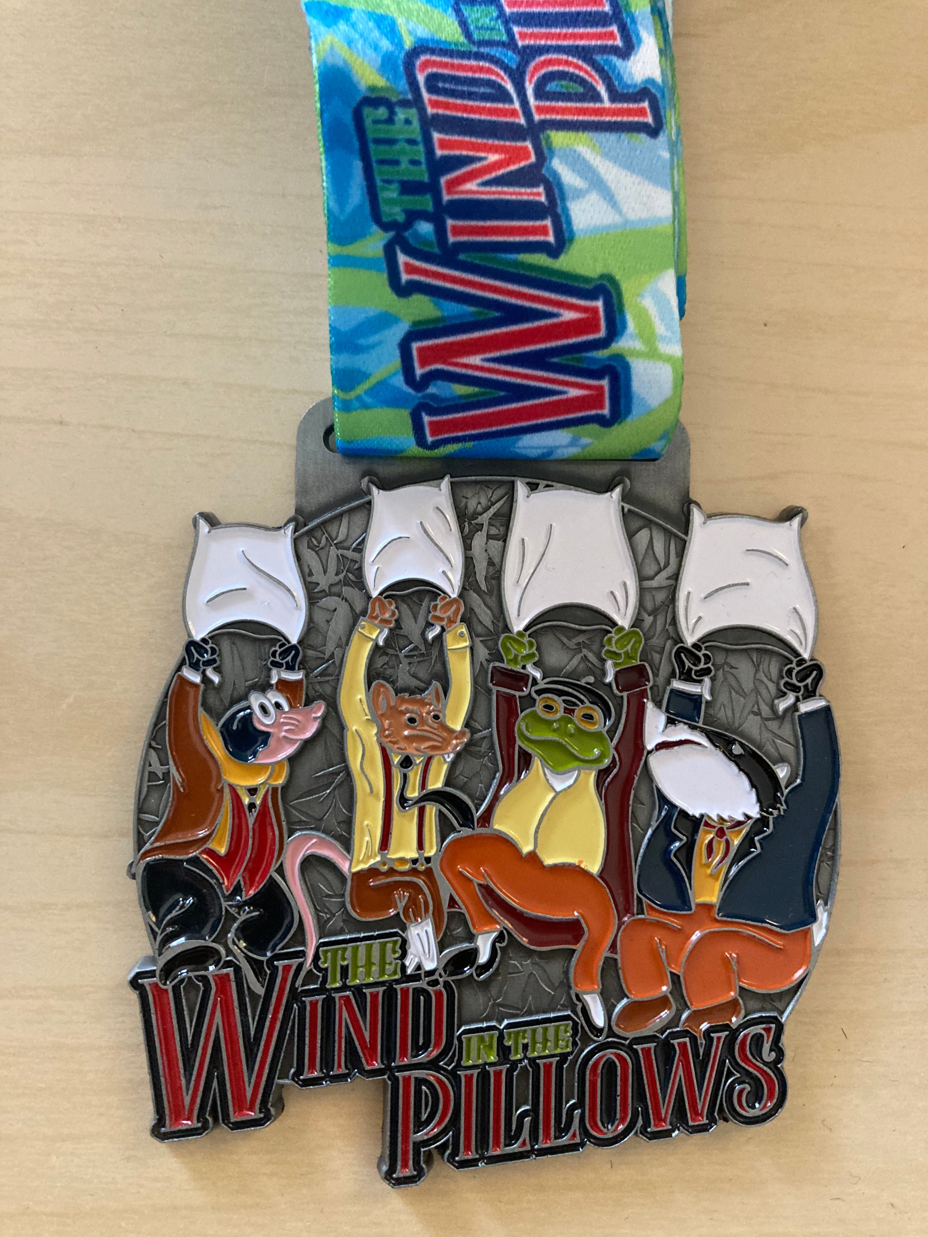 A medal with colorful cartoon characters of a rat, a mole, a frog, and a toad, all holding pillows above their heads. The lanyard connected to the medal has "The Wind in the Pillows" written on it in bold red letters with a blue and green background.