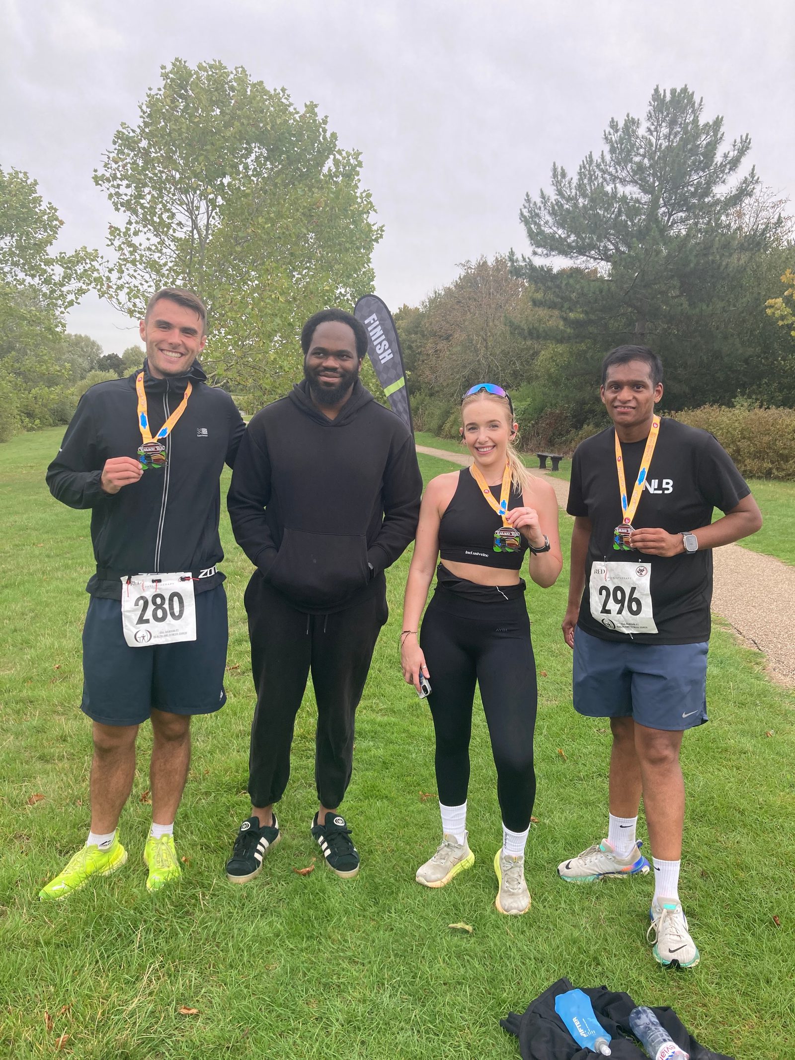 Four people stand on grass outdoors, three wearing athletic gear and race numbers with medals, smiling after a race. A "Finish" flag is in the background along with trees and a cloudy sky.