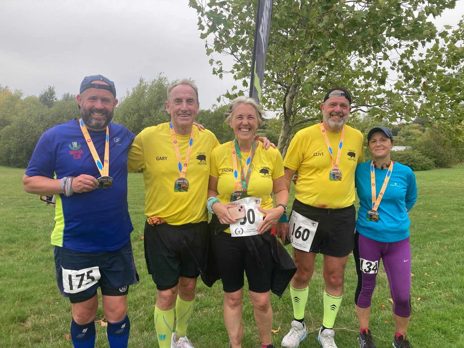 Five smiling runners stand outside on grass wearing race bibs and medals. Four wear yellow shirts, one wears blue, and one wears a blue cap. Trees and cloudy sky are in the background.