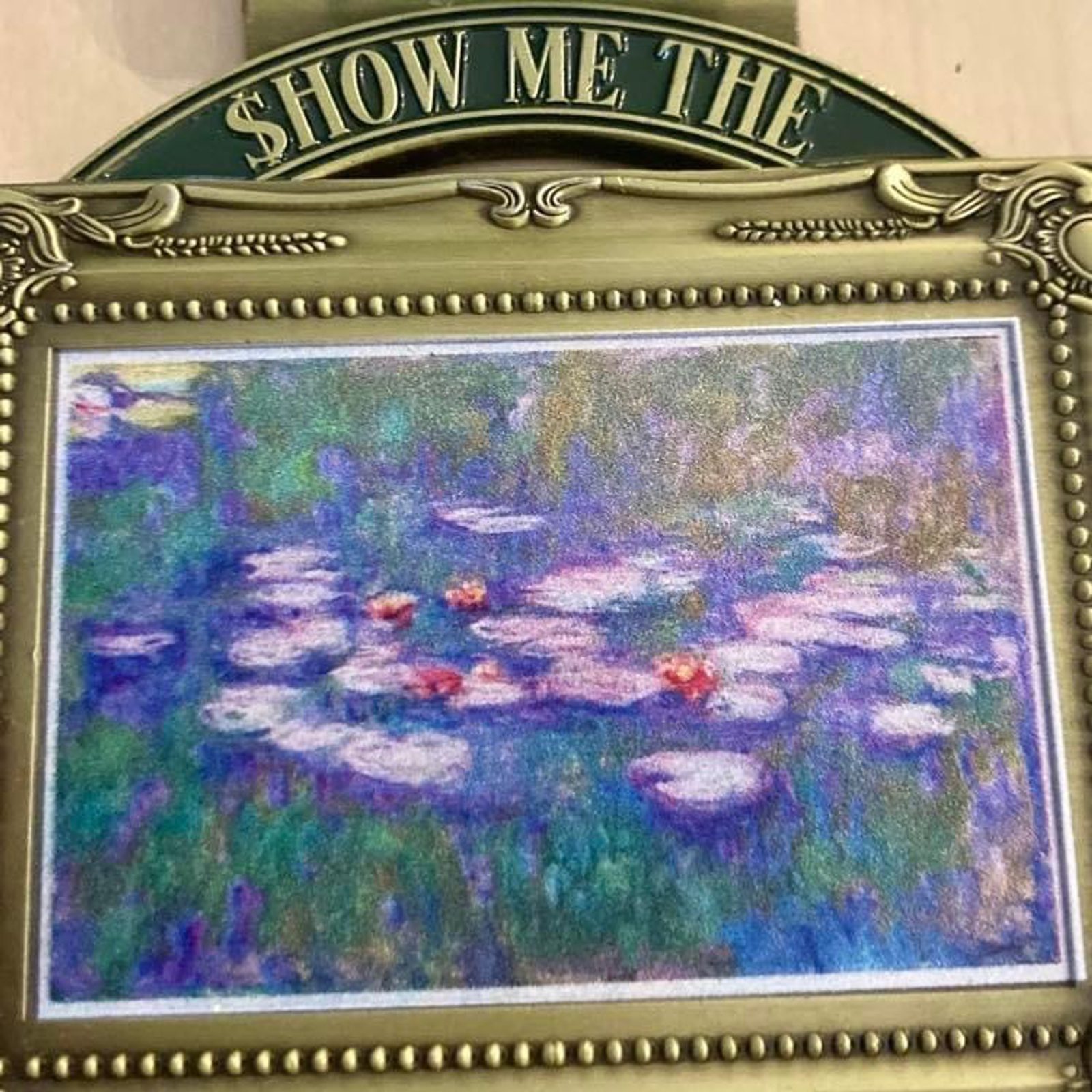 Framed impressionist painting of water lilies with a metallic decorative border. At the top, text reads "SHOW ME THE $" indicating a playful take on valuing art. The painting features vibrant colors, including shades of purple, blue, green, pink, and white.