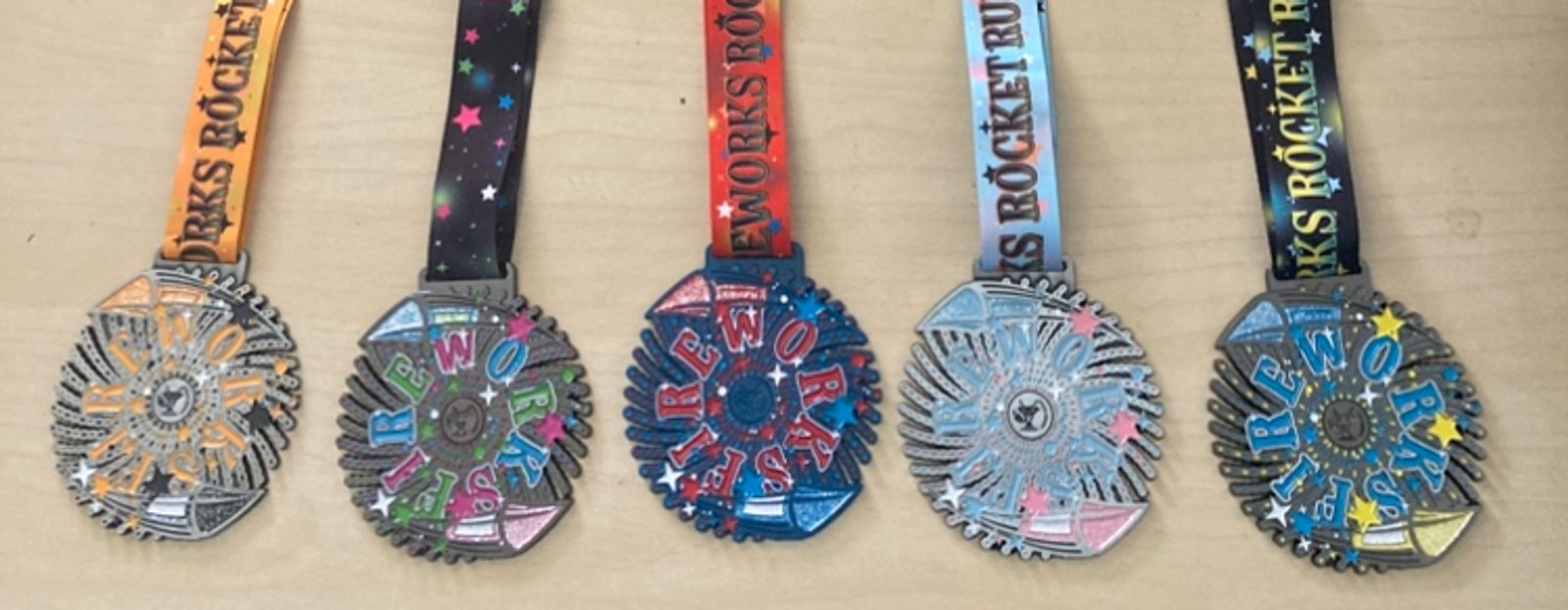 Five colorful medals with star-patterned ribbons, each reading “Fireworks Rocket Run,” are arranged in a row on a light wood surface. The medals have a fireworks design and different colored accents.