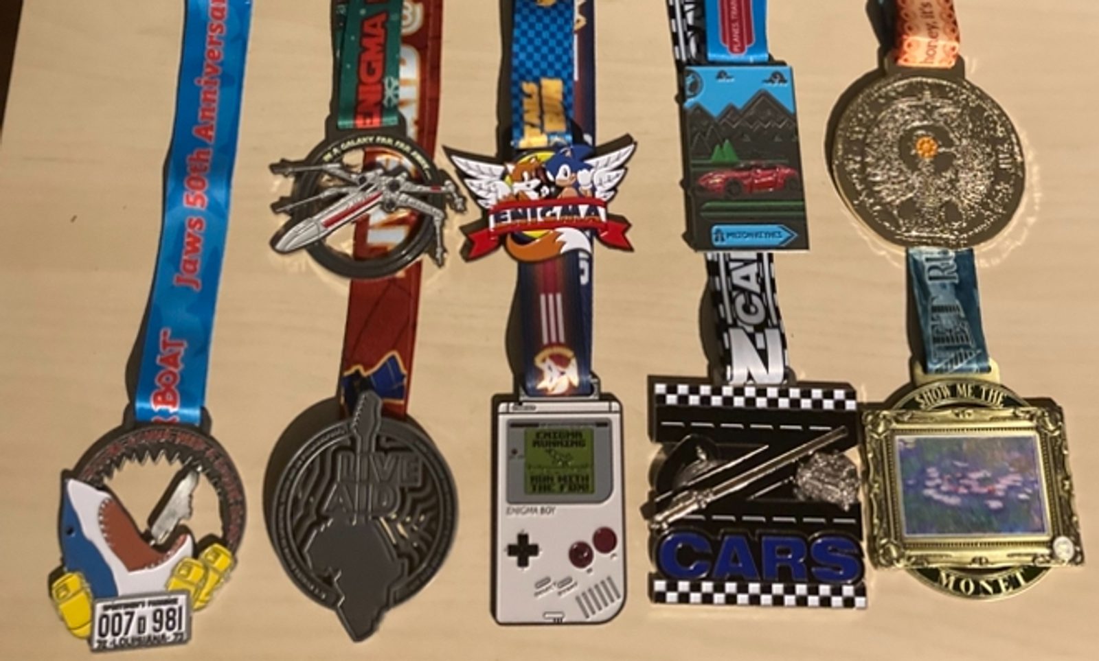 Nine colorful race medals with various shapes and designs, including images of airplanes, cars, a shark, a mountain, a Game Boy, and a circular gold medallion, are displayed on a light wooden surface.