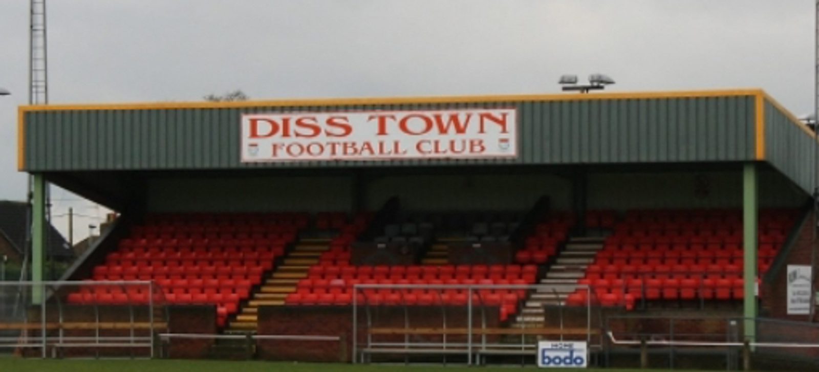 A football stadium stand with red seats and a sign reading "DISS TOWN FOOTBALL CLUB" above the seating area. The stand is partially covered with a green and yellow roof.