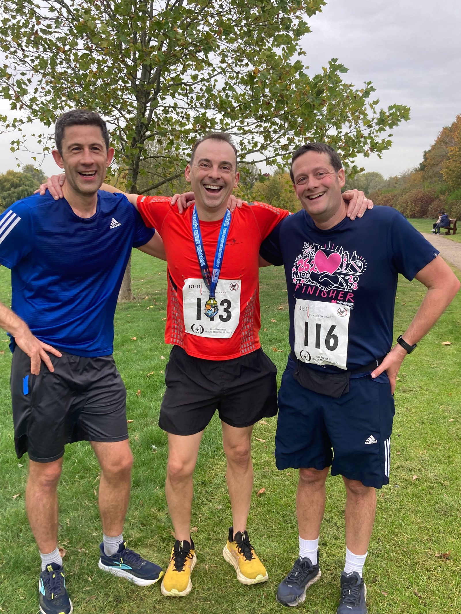Three men stand smiling with their arms around each other after a race, all wearing athletic clothes and numbered race bibs. The man in the center wears a medal. Trees and grass are visible in the background.