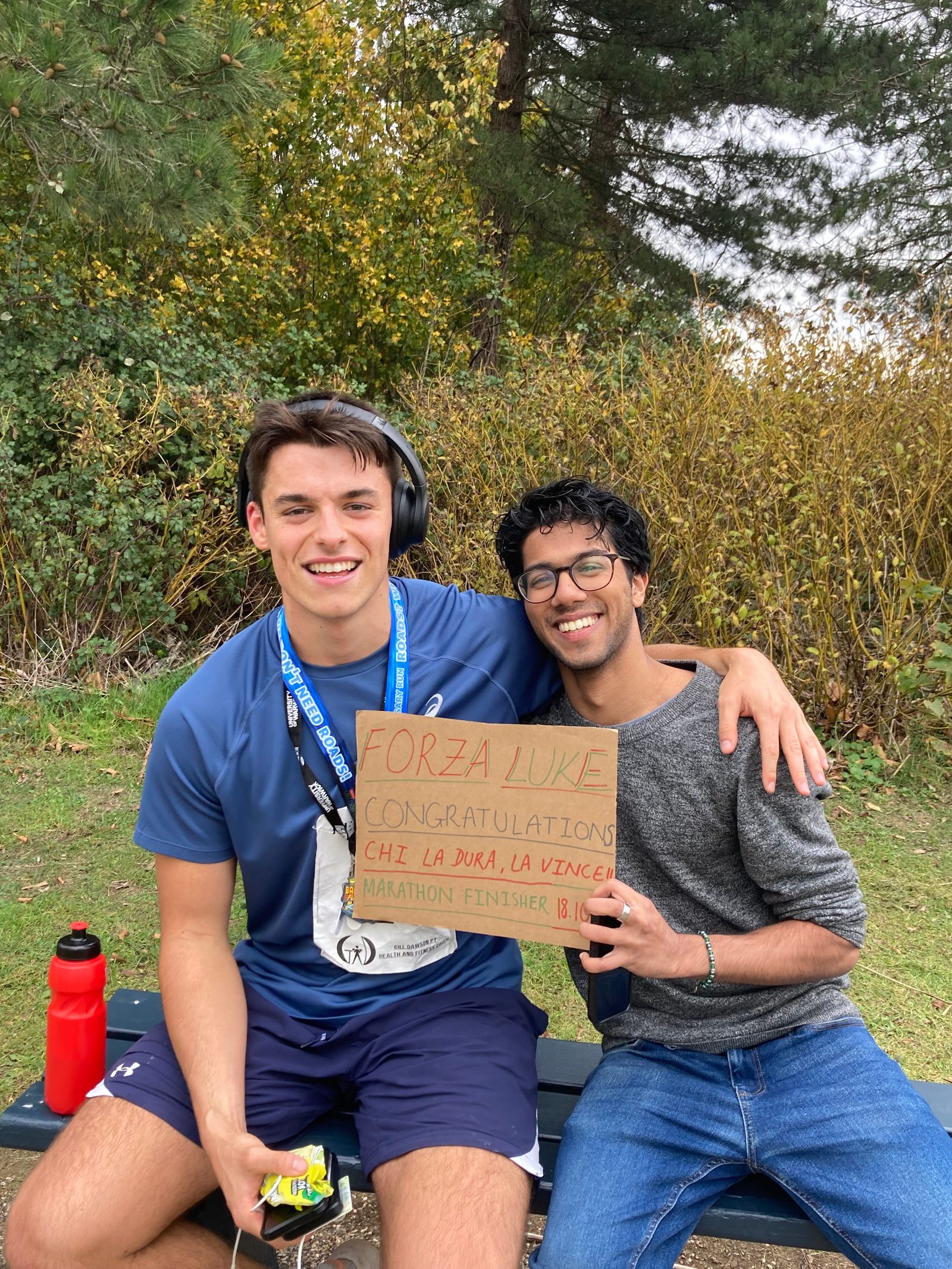 Two young men sit on a bench outside, one wearing a marathon medal and headphones, smiling with his arm around the other, who is holding a congratulatory sign that reads "FORZA LUKE" and other handwritten text. Trees and bushes are in the background.