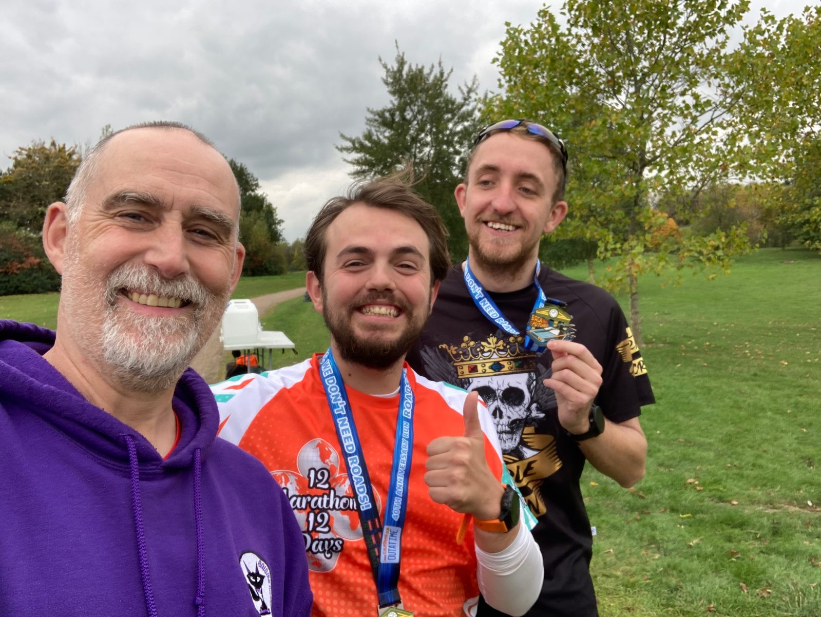 Three smiling men stand outdoors on grass, wearing athletic clothes and medals. One man in a purple hoodie takes a selfie; the other two wear running shirts and proudly display their medals. Trees and cloudy sky are in the background.