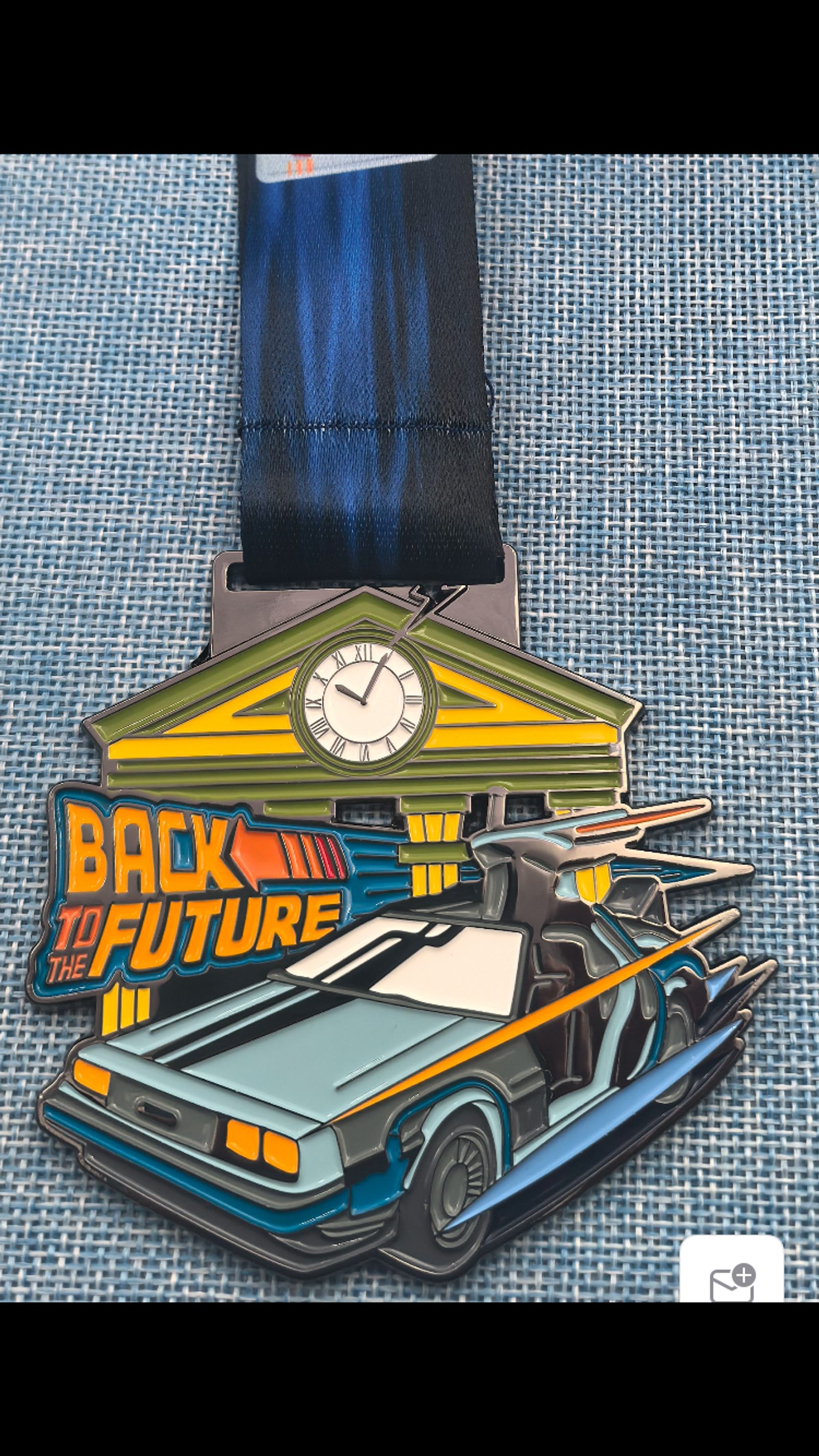 A medal featuring the DeLorean time machine from "Back to the Future," with the Hill Valley courthouse and clock in the background, attached to a blue ribbon, resting on a textured blue surface.