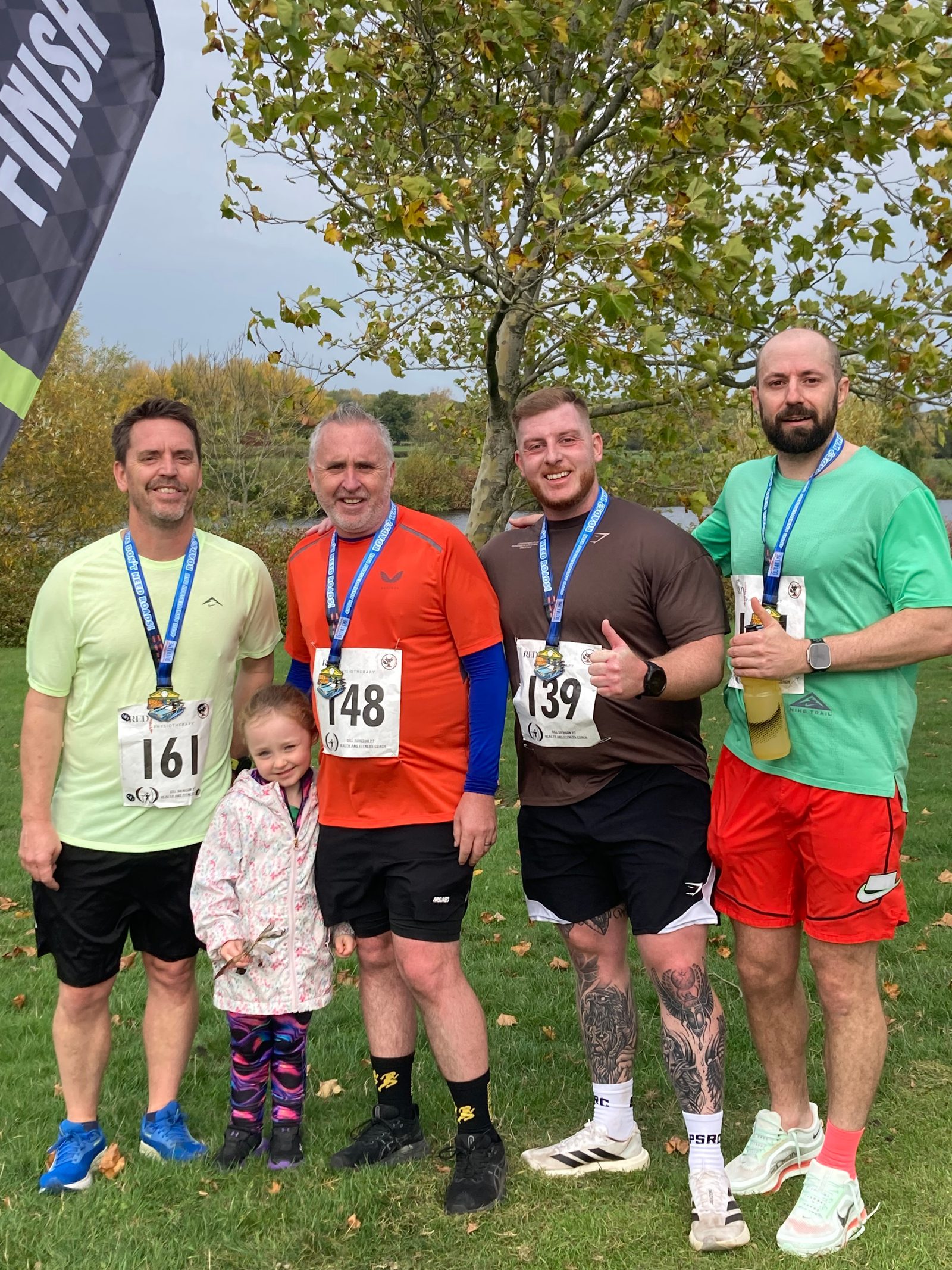 Four men wearing race bibs and medals stand together outdoors with a young girl in front. They are smiling, dressed in athletic clothes, and a "Finish" flag and leafy tree are visible in the background.
