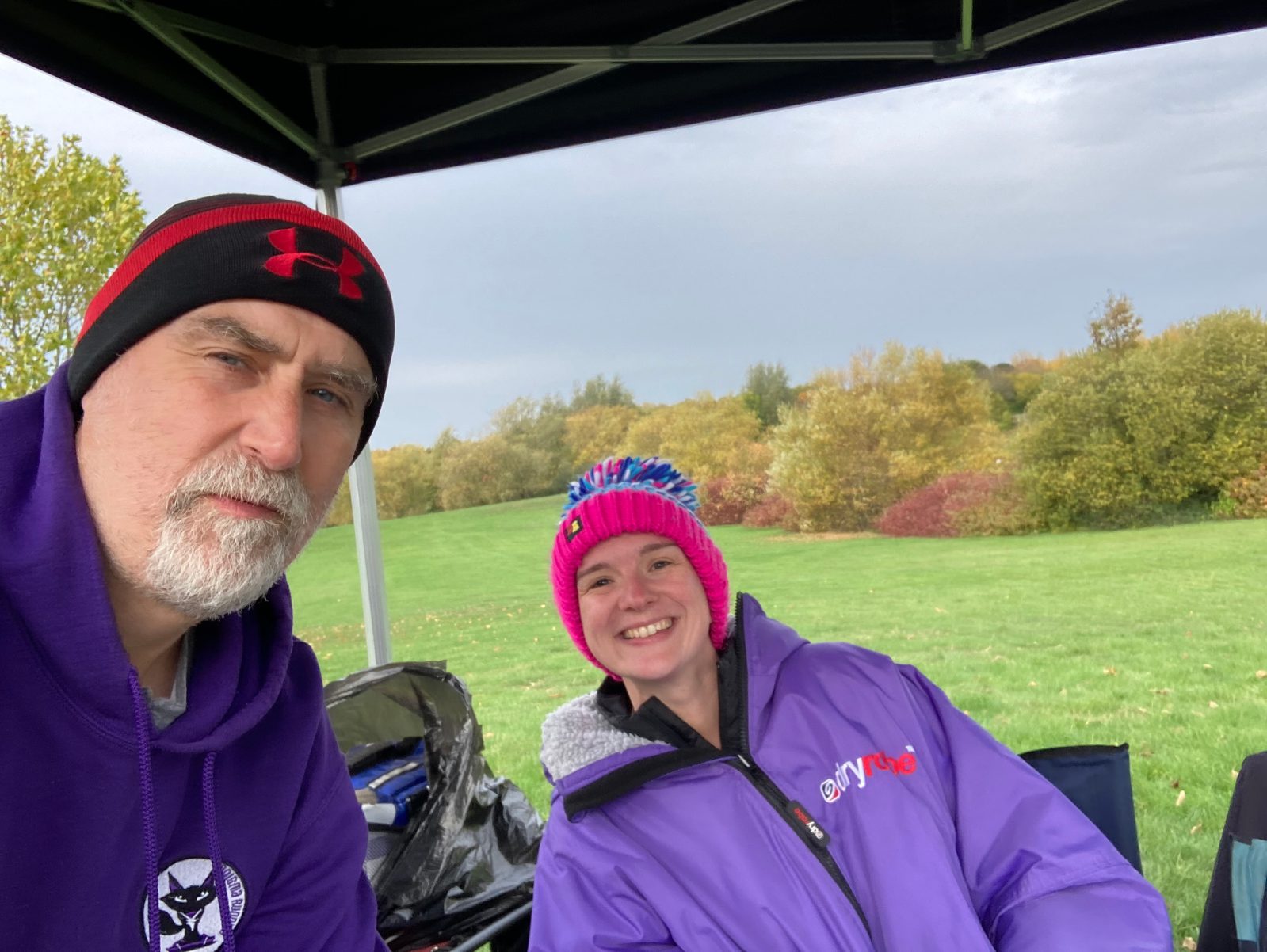 A man in a purple hoodie and black-and-red beanie and a smiling young person in a purple jacket and colorful knit hat sit under a canopy in a grassy park with autumn trees in the background.