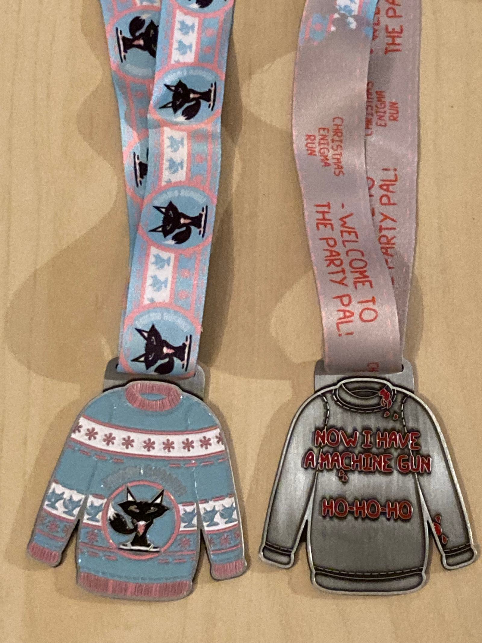 Two medals shaped like Christmas sweaters. The left medal is blue with a cat design on the ribbon. The right medal has "Now I have a machine gun, Ho-Ho-Ho" written on it, with a pink ribbon featuring text about a Christmas event.