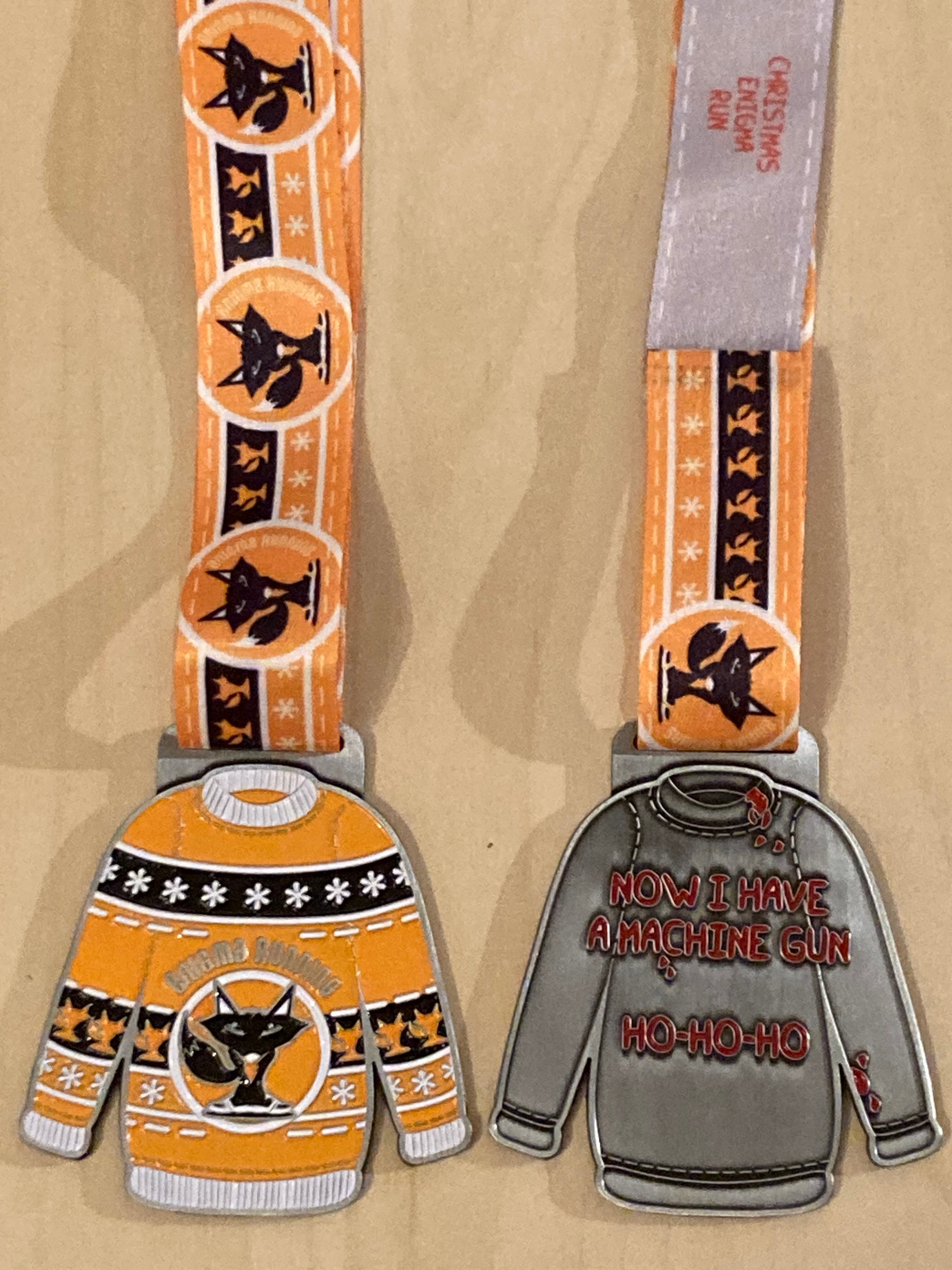 Two decorative Christmas-themed medals with ribbons are displayed. The left medal has an orange sweater with black cats and festive patterns, while the right medal has a grey sweater with "Now I have a machine gun. Ho-ho-ho" in red letters.