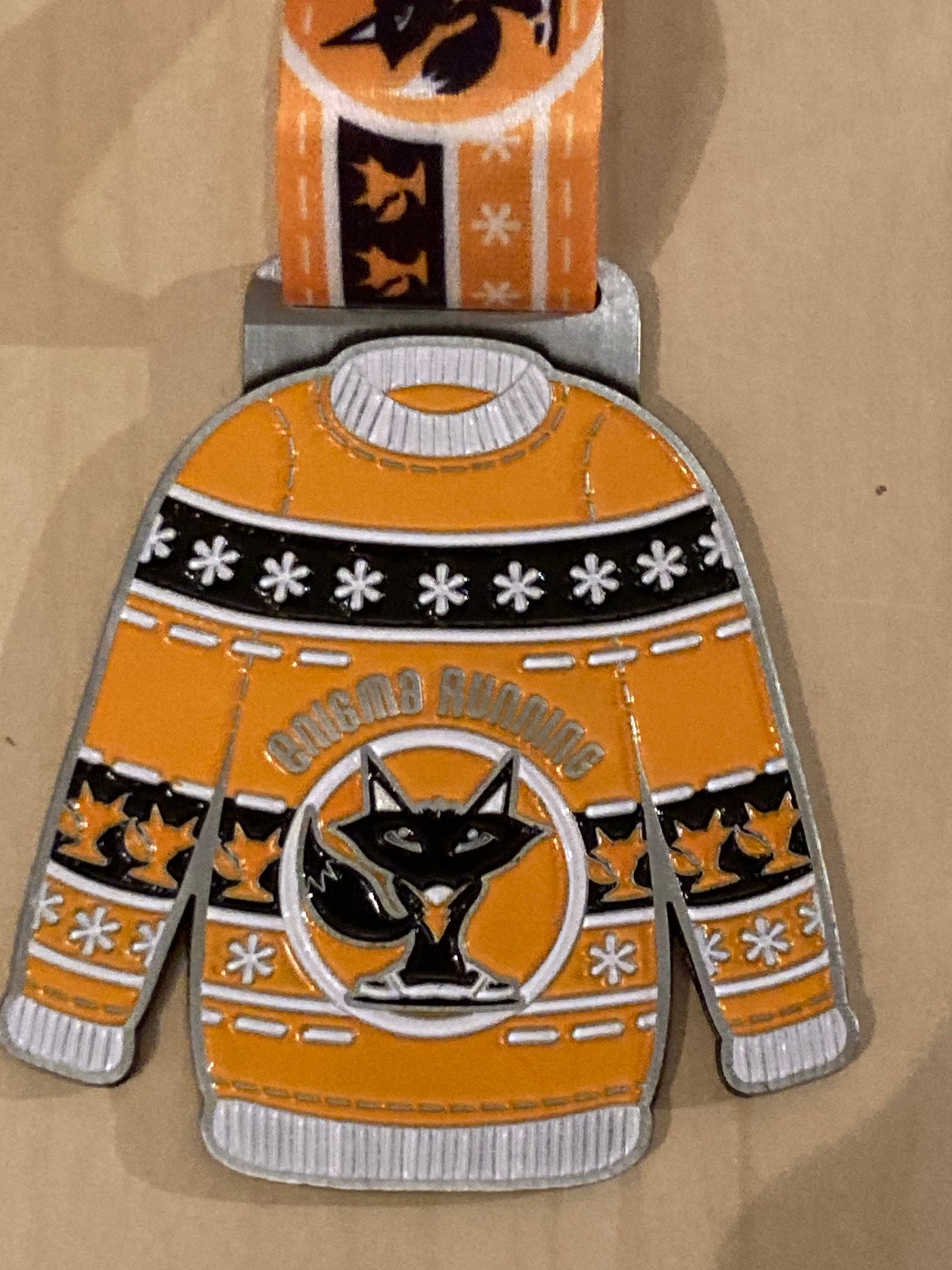A festive medal with a design of an orange winter sweater featuring snowflakes, a black and white fox, and the words "RUNNING" above it. The ribbon has a matching pattern with fox silhouettes and snowflakes.