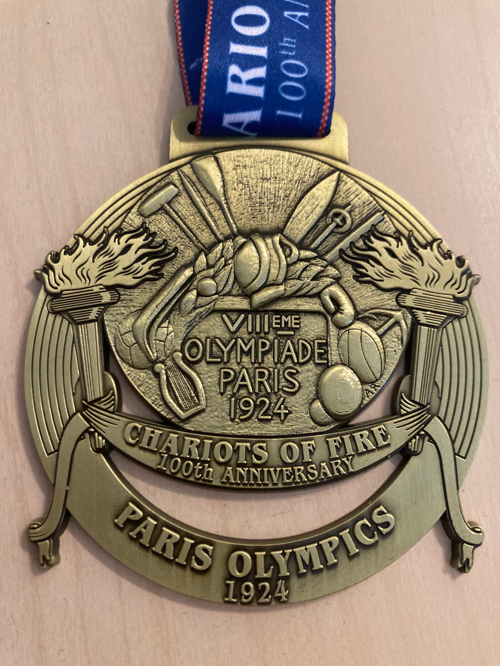 A gold medal with an image of two torches and various sports equipment, labeled "VIIIe OLYMPIADE PARIS 1924,” "CHARIOTS OF FIRE 100th ANNIVERSARY," and “PARIS OLYMPICS 1924.” The medal hangs from a blue ribbon with "MARION DAVIES 100th ANNIVERSARY" written on it.