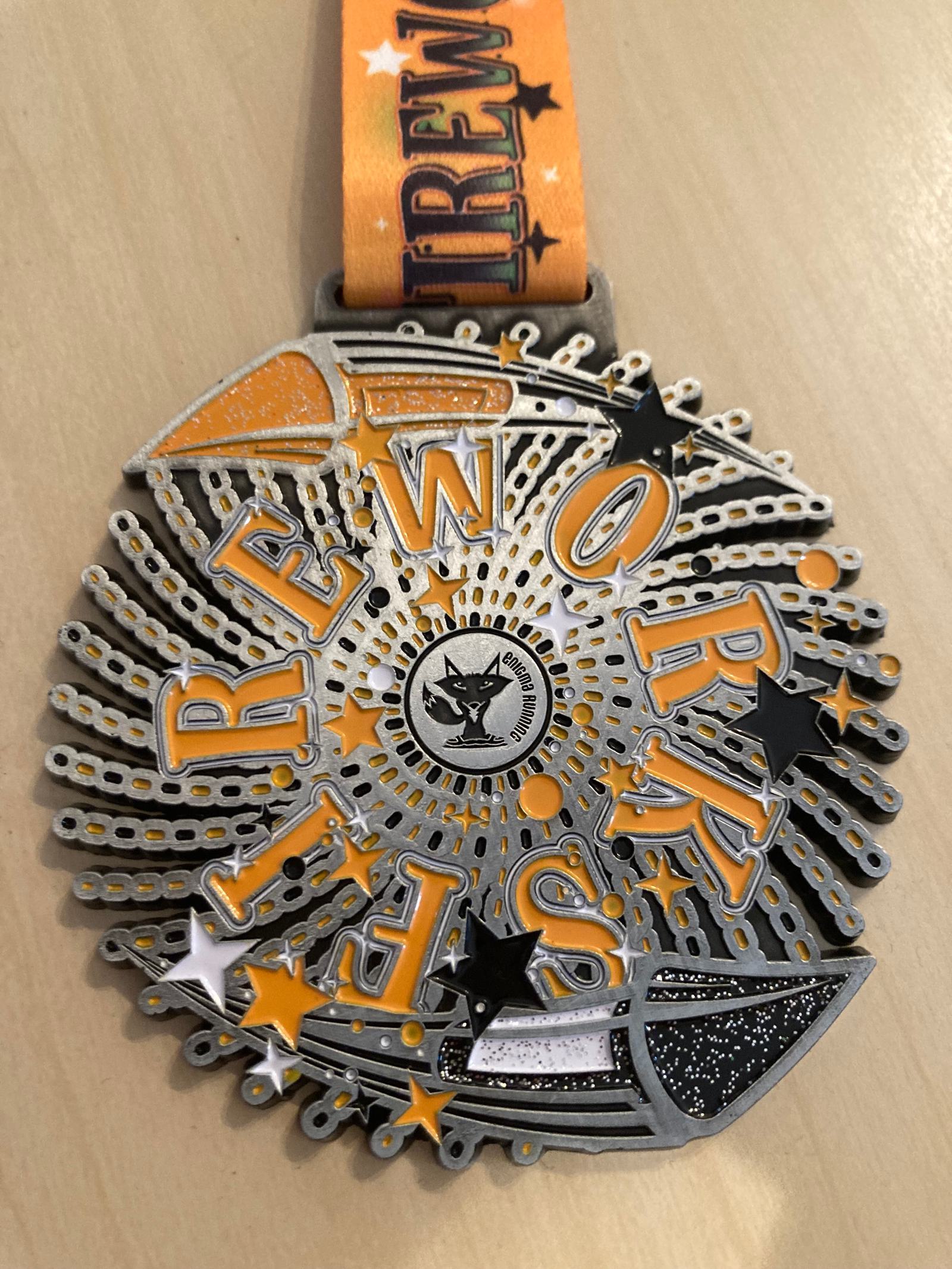 A silver medal with the words "Fireworks" in orange and black text. It features star designs and a central cat logo. The medal hangs from an orange ribbon with the same word repeated.