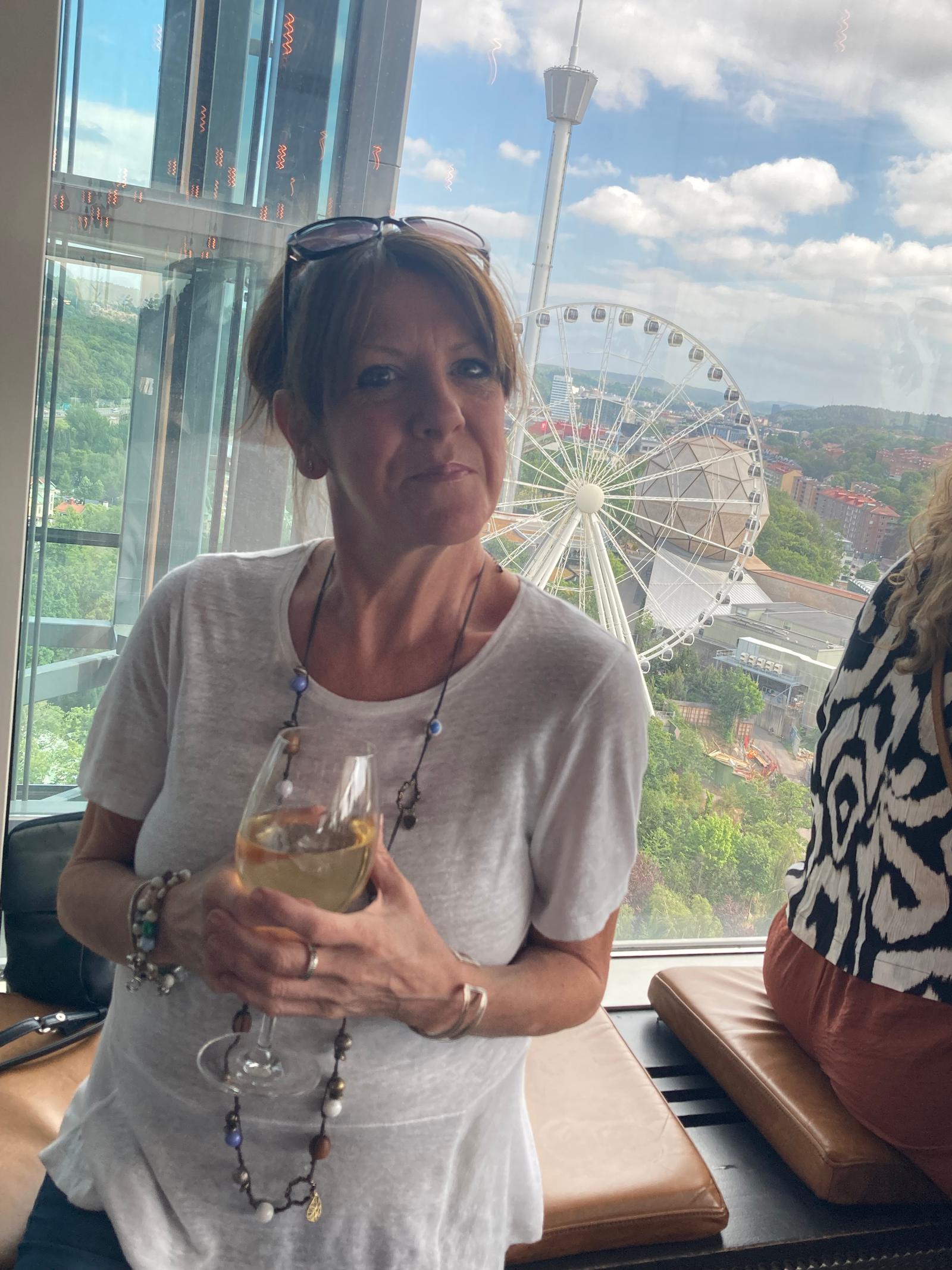 A woman wearing a white shirt and a beaded necklace holds a glass of wine with both hands. She is standing in front of a large window, with a ferris wheel visible in the background on a bright, sunny day. She has sunglasses resting on her head.