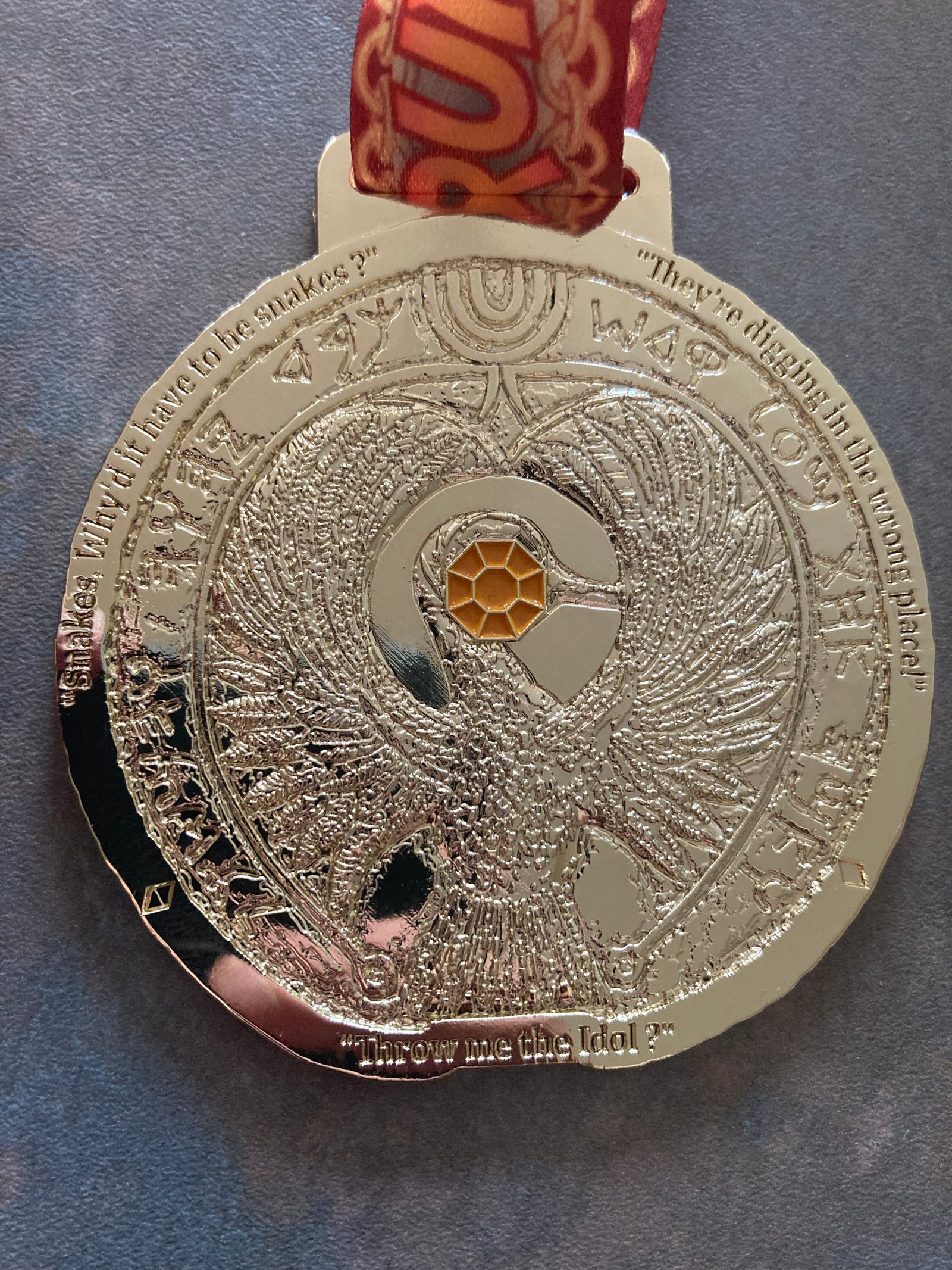 A close-up photo of a silver medal with intricate embossed designs, including a bird with outstretched wings, text encircling the bird in a circular pattern with some in an unknown script, and a yellow honeycomb-shaped center. The medal is attached to a red, patterned ribbon.