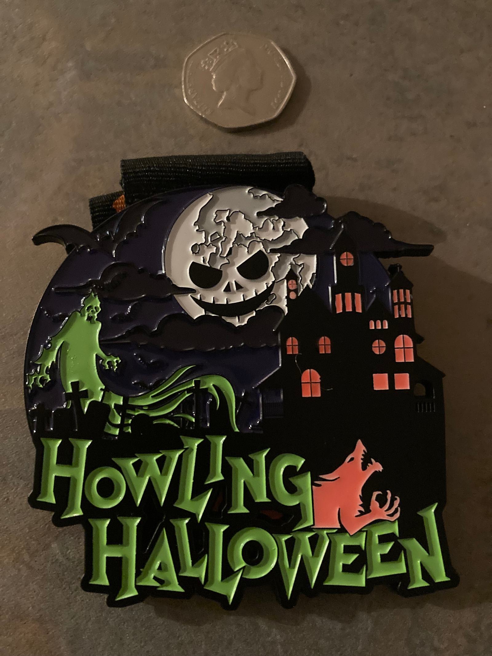 A decorative Halloween-themed medal featuring a spooky moon with a face, a haunted house, and a howling wolf. The text "Howling Halloween" is prominently displayed at the bottom. A British 50p coin is placed above for size reference.