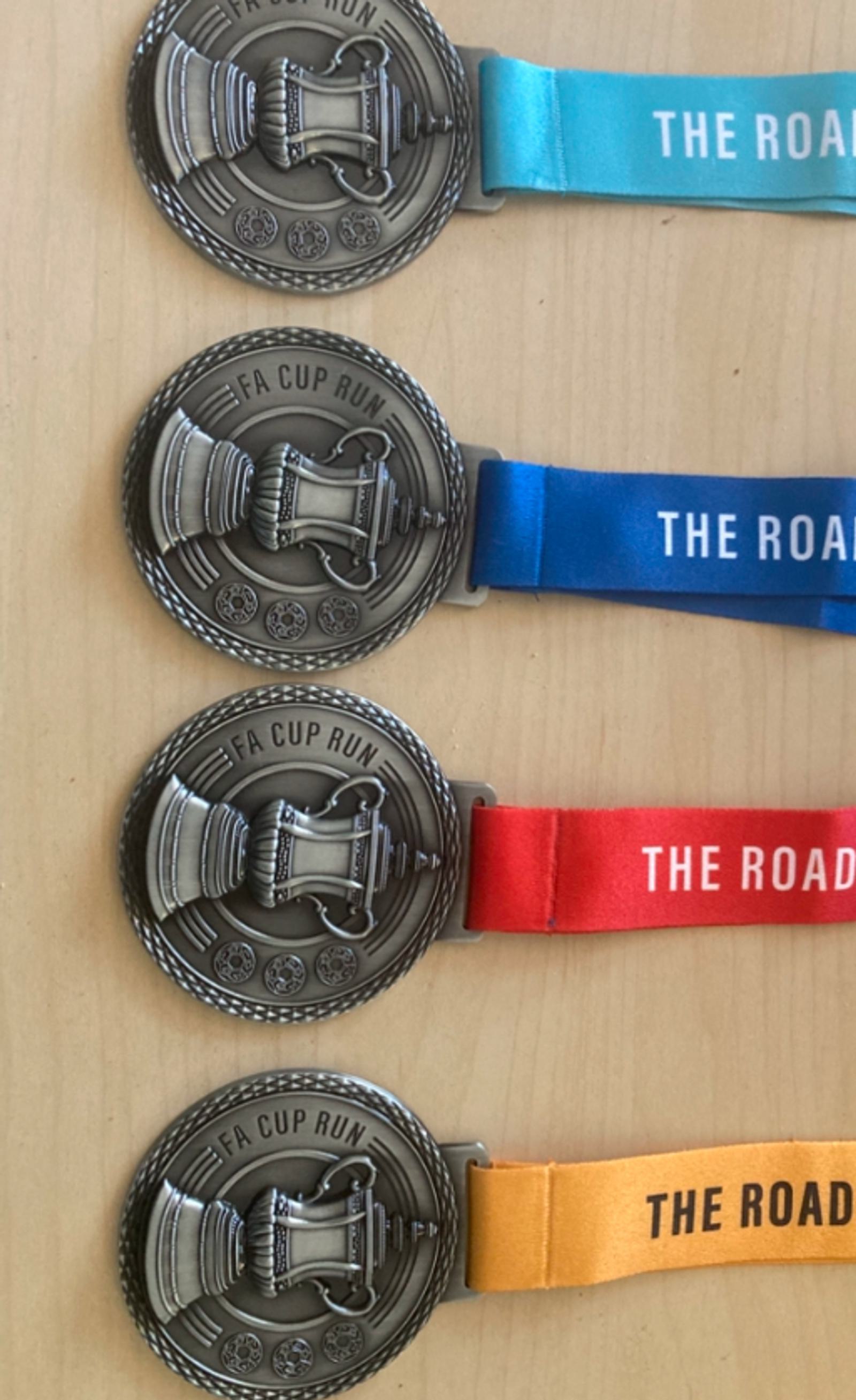 Four FA Cup Run medals are displayed on a wooden surface. The medals feature an image of a cup and are attached to different colored ribbons: two blue, one red, and one orange.