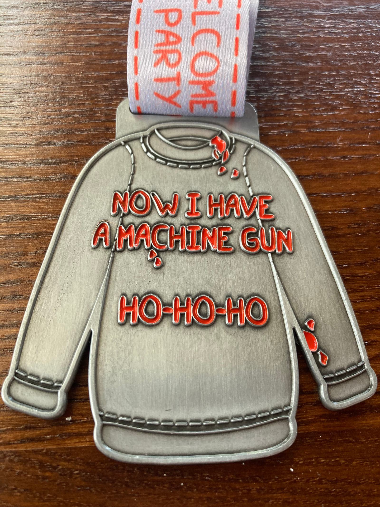 A metal Christmas ornament in the shape of a sweater has the text "NOW I HAVE A MACHINE GUN HO-HO-HO" engraved in red letters on its front. The ornament is attached to a ribbon that has a partial, unclear design and text. The background is a wooden surface.