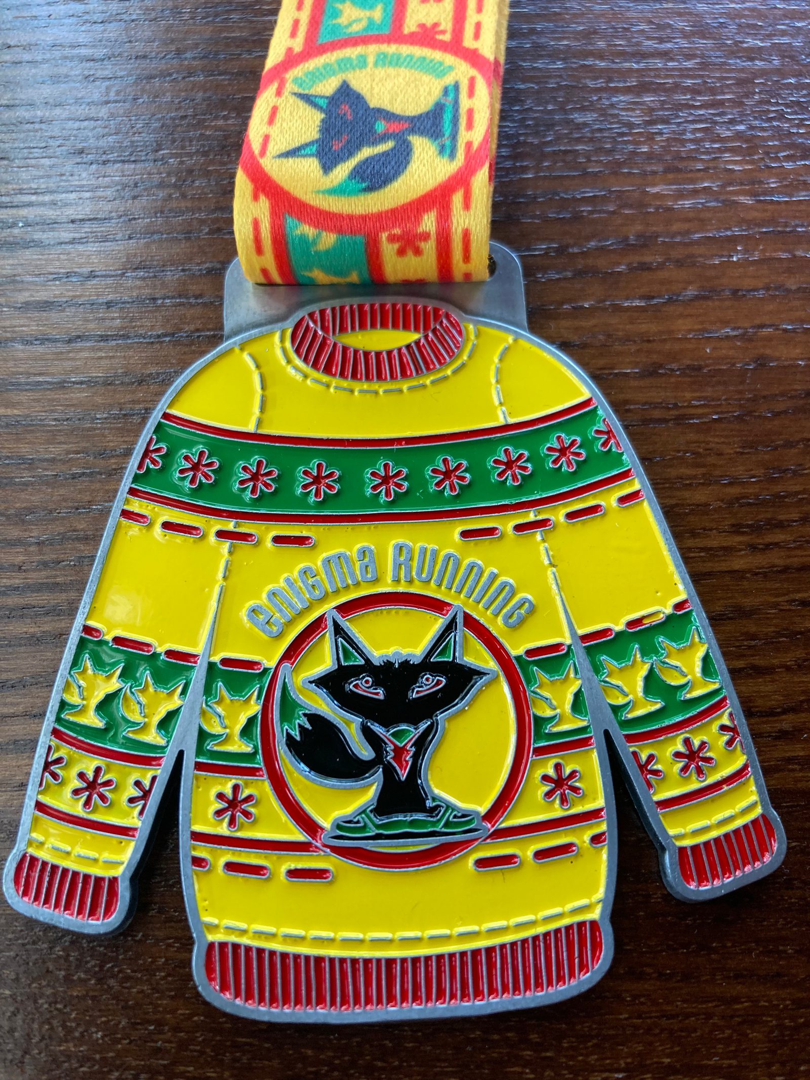 A colorful race medal shaped like a sweater rests on a wooden surface. The sweater is yellow with red and green festive designs, featuring a black fox in the center. "Enigma Running" is written above the fox. The attached ribbon has a repeating fox pattern.