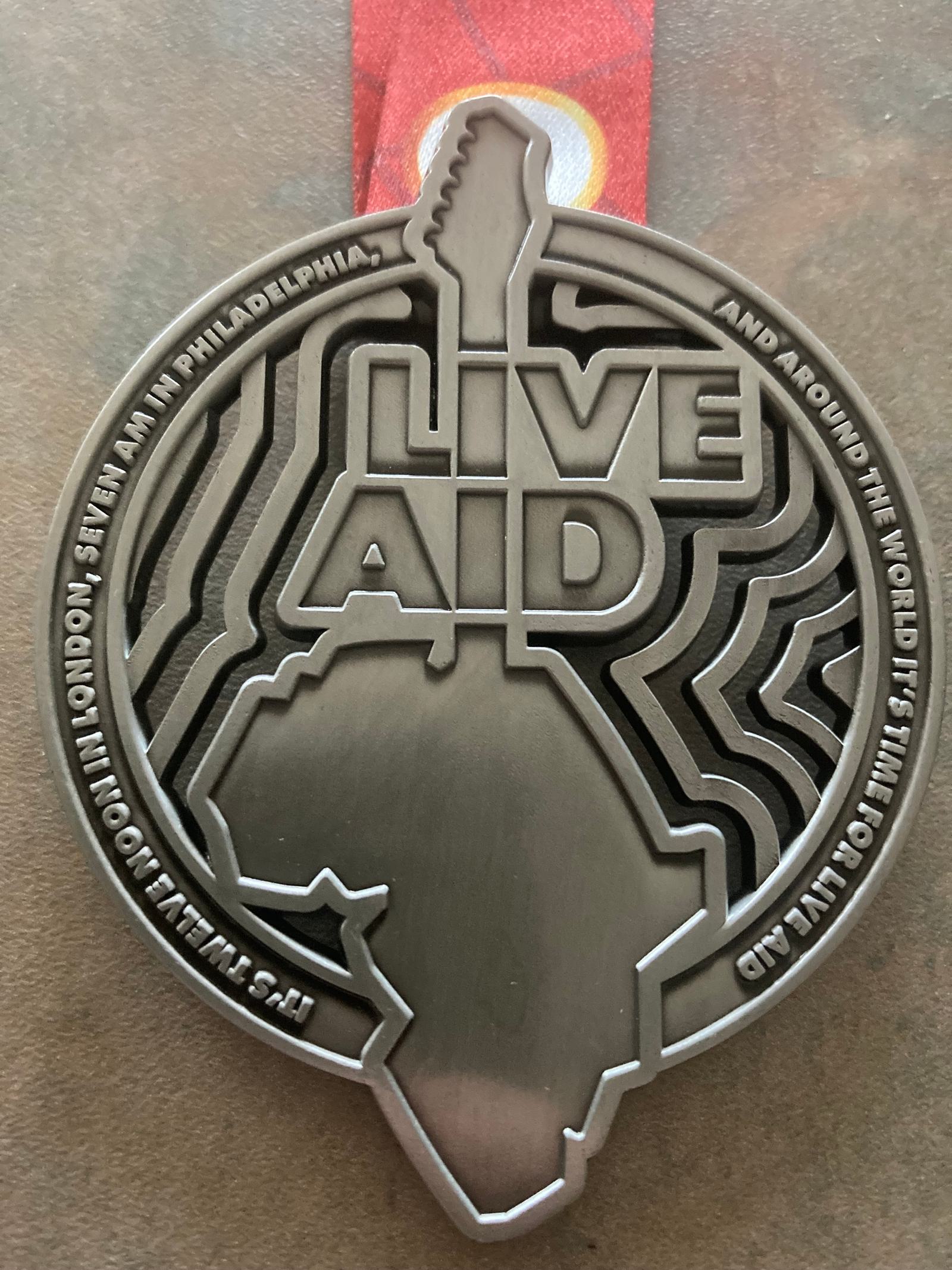 A round, silver medal with the words "Live Aid" and a guitar silhouette engraved above an outline of Africa. Text around the edge reads, "It's time to help in Philadelphia and around the world, it's time for Live Aid in London.