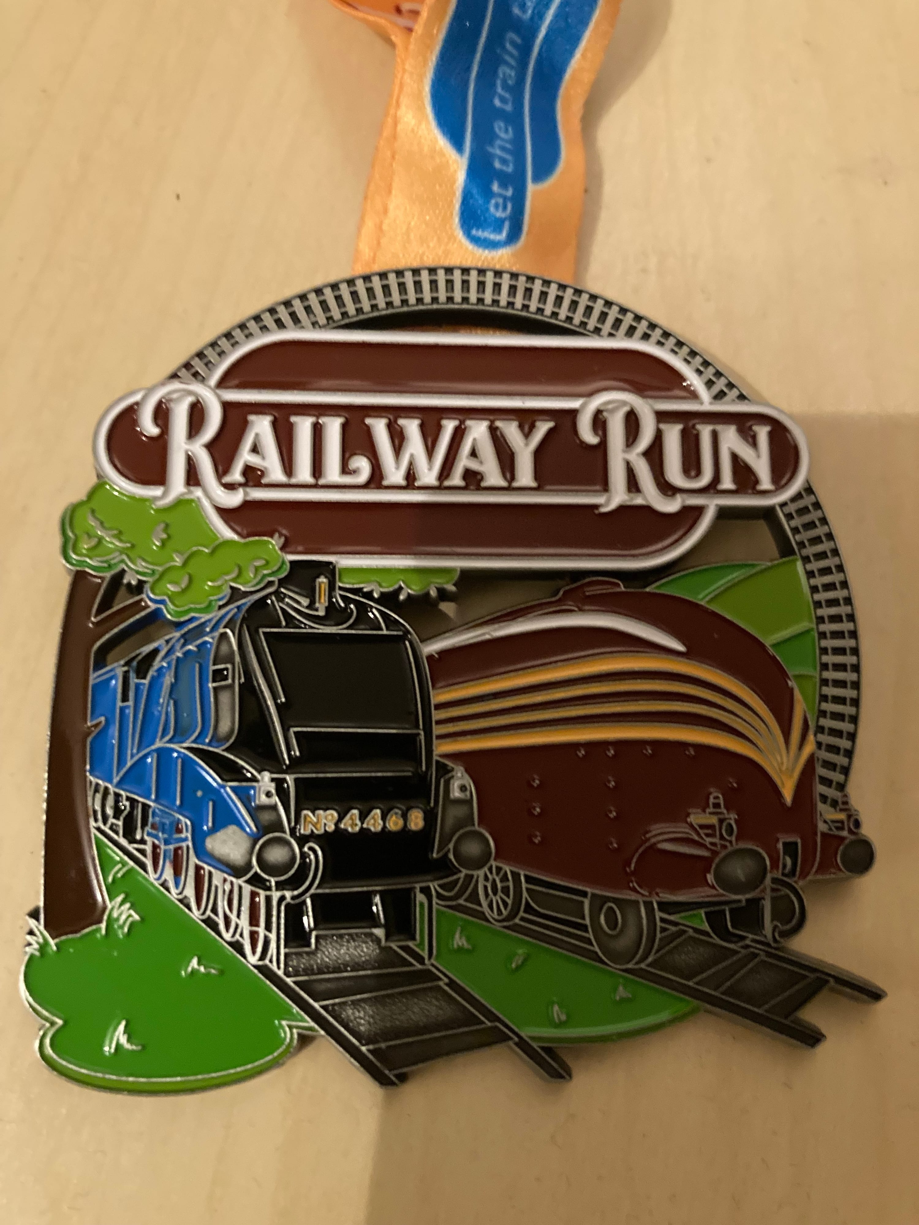 A medal with two classic trains on tracks under the text “Railway Run.” One train is blue and the other is maroon. The medal has a ribbon attached and is set on a light-colored surface.
