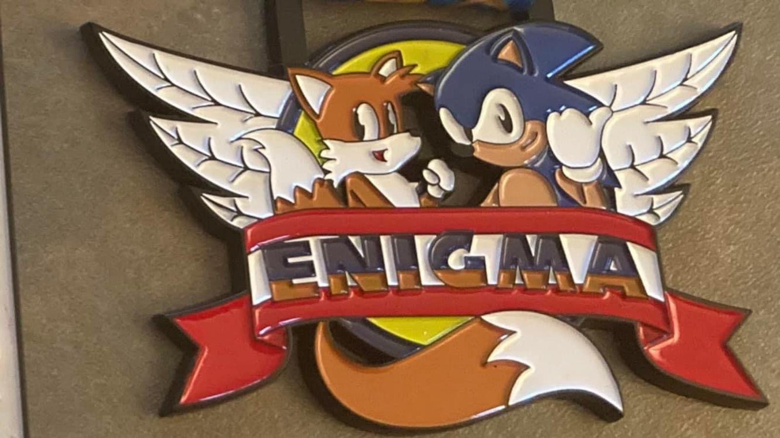 A sign featuring two winged cartoon characters, one orange with white tails and the other blue with spikes, facing each other. The characters are framed by a banner with the word "Enigma" written on it in bold letters.