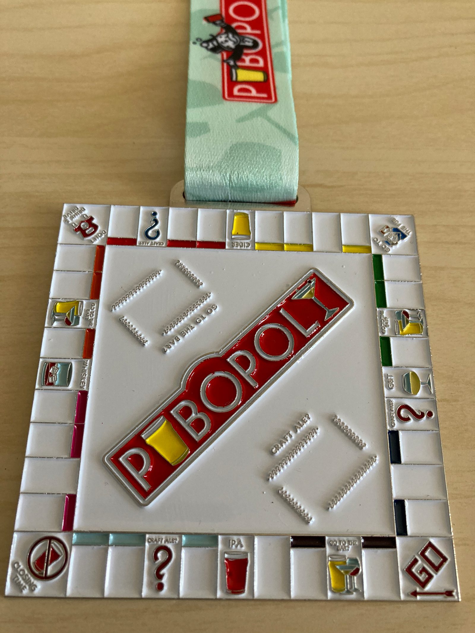 A square medal designed to resemble a Monopoly game board. The center features the text "Pubopoly" in red and white. The medal is attached to a light green ribbon. The board edges have game spaces illustrated with various drink icons.