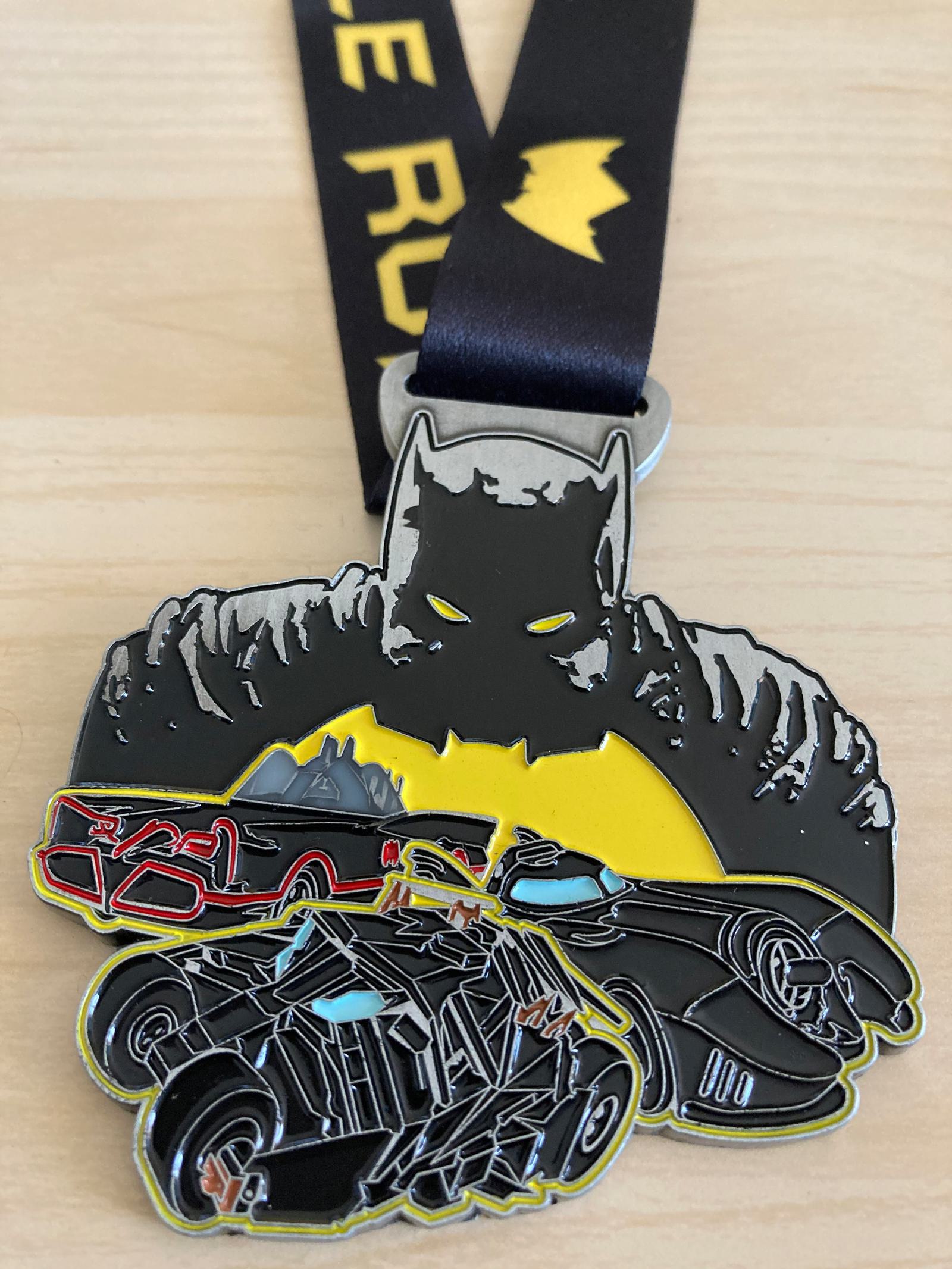 A close-up view of a Batman-themed medal placed on a wooden surface. The medal features an image of Batman with a yellow bat symbol behind him, and illustrations of three Batmobiles at the bottom. A black ribbon with a yellow emblem is attached to the medal.