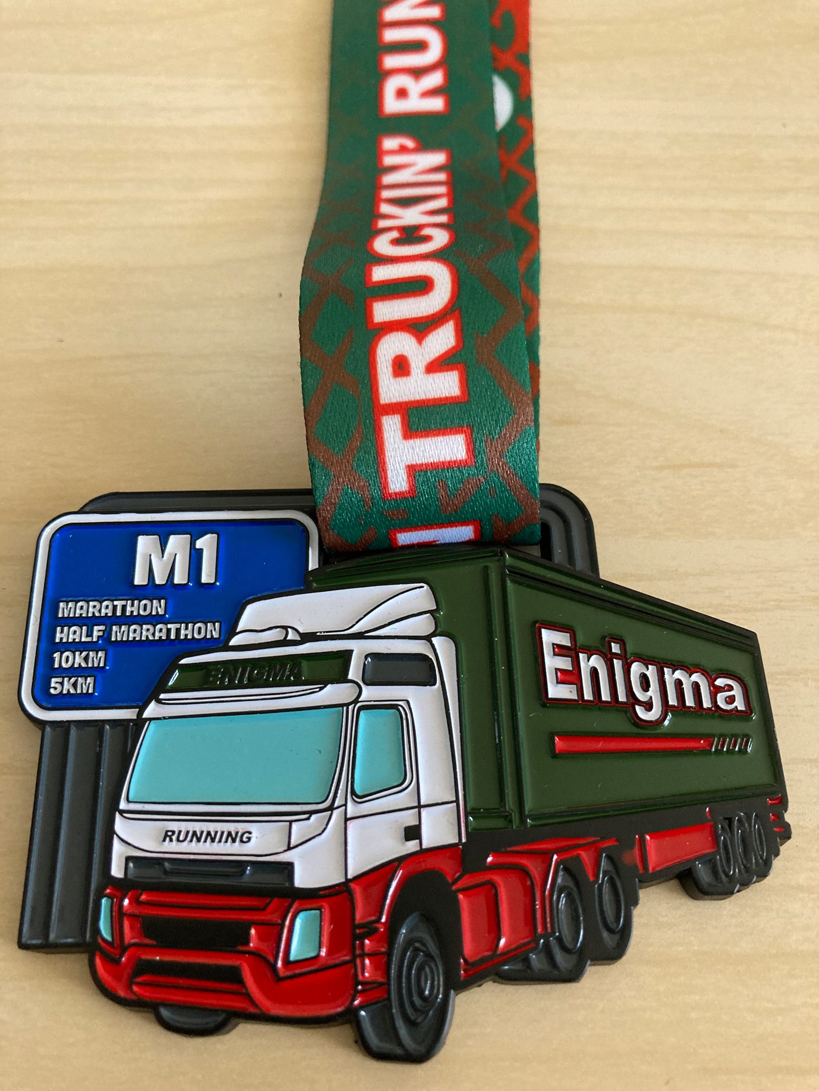 A medal depicting a white truck with a green trailer that reads "Enigma." The ribbon is green with red highlights, displaying the text "TRUCKIN' RUNNING." A sign stays beside the truck with text: "M1," "Marathon," "Half Marathon," "10KM," and "5KM.