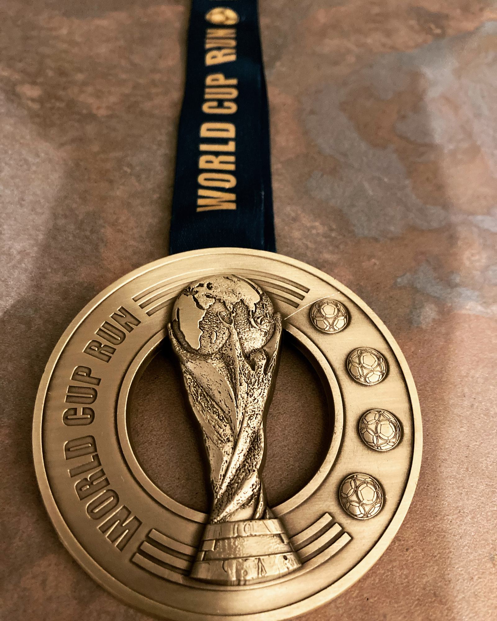 A gold medal with "WORLD CUP RUN" inscribed along the bottom and on the surrounding navy ribbon. The center features a raised image of the FIFA World Cup trophy. Three soccer balls are etched in a vertical line on the right side of the medal.