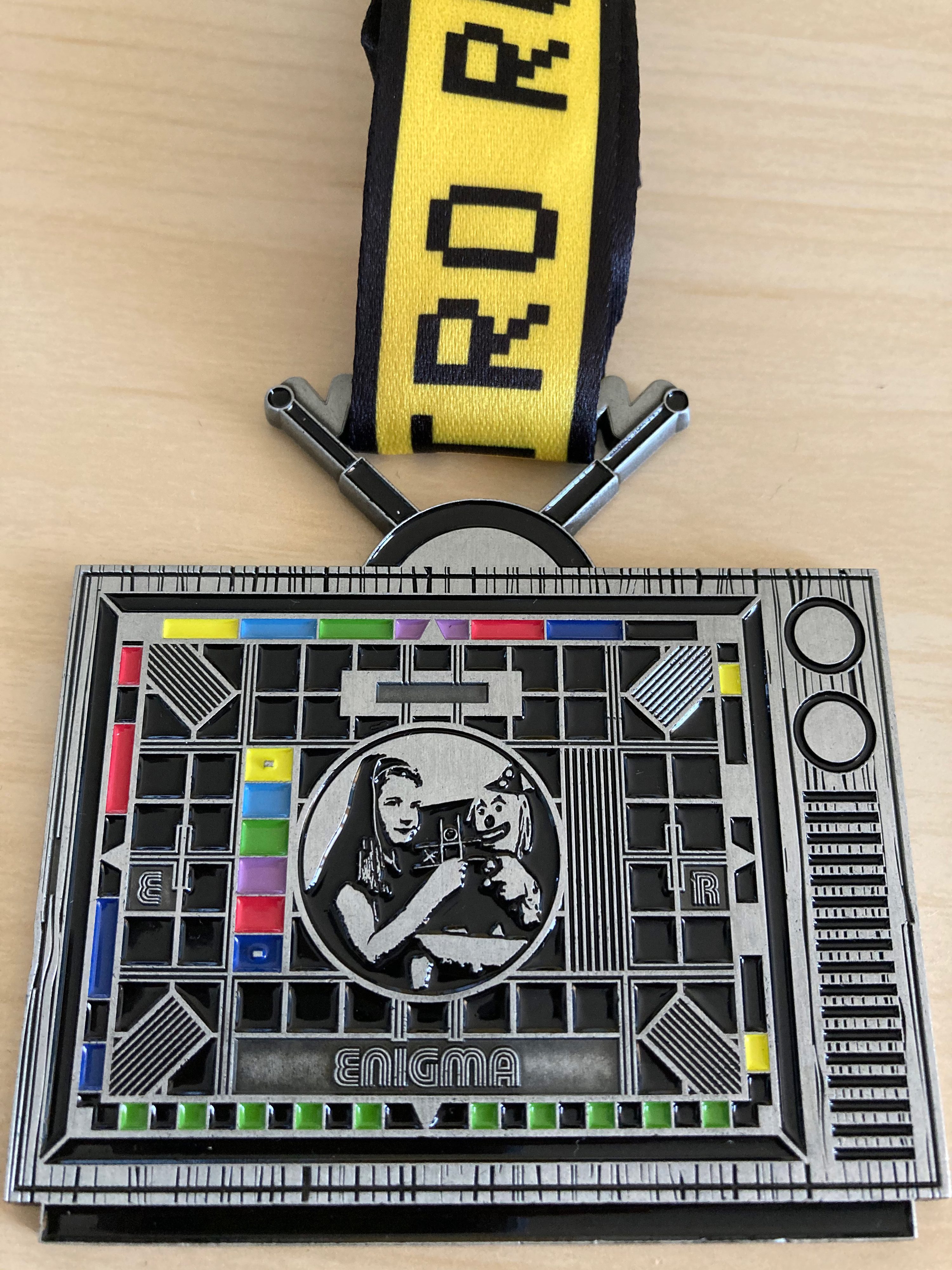 A rectangular metallic medal designed to resemble a retro TV set with colorful buttons and a central circular image of two people. The word "ENIGMA" is engraved at the bottom. The medal hangs from a yellow and black ribbon with text and symbols.