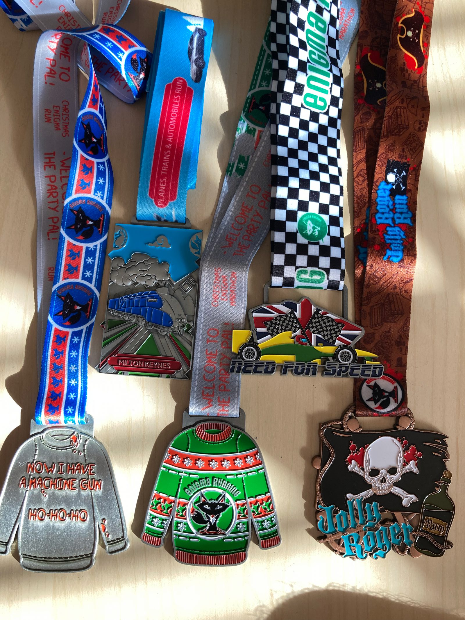 A collection of six colorful medals with various designs on a light wooden surface. The medals have themes like running, holiday sweaters, and racing, featuring festive elements, skulls, and cars. Each medal hangs from a ribbon with coordinating graphics.
