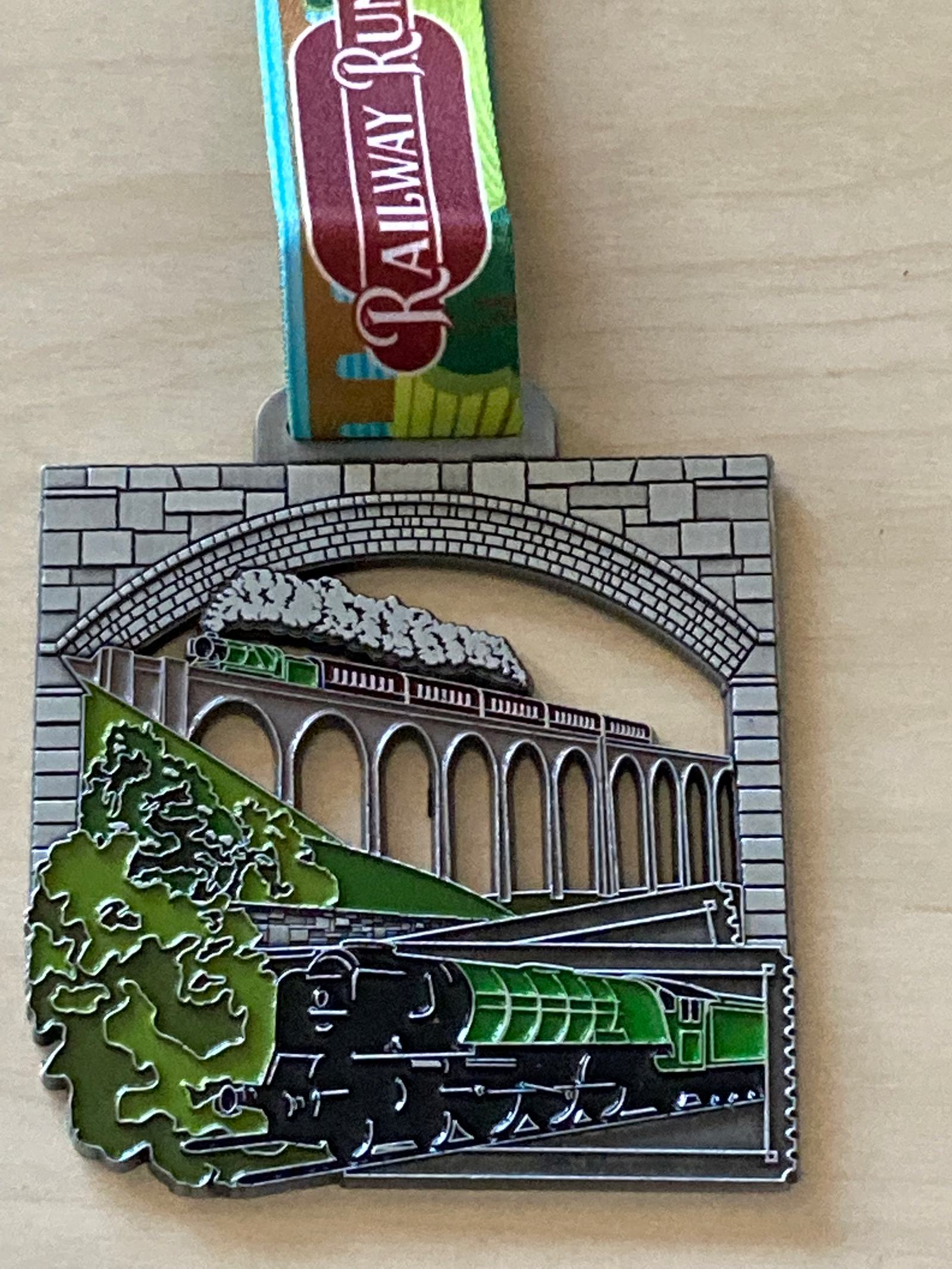 A square-shaped medal with a colorful ribbon depicts a green steam train crossing a viaduct bridge. The background features stone brick arches and green trees. The ribbon, attached to the top of the medal, is labeled "Railway Run.